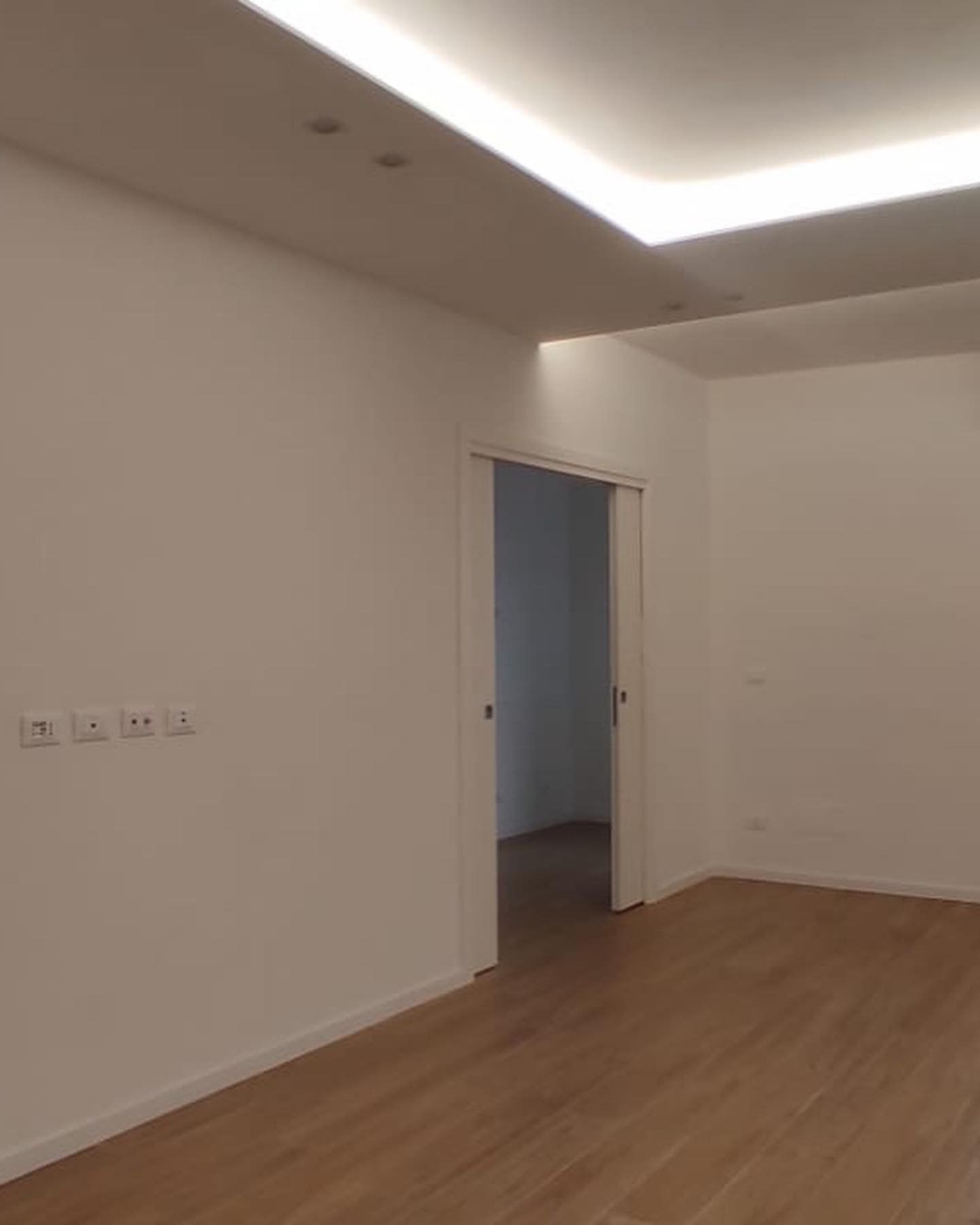 Stanza vuota con pavimenti in legno chiaro, pareti bianche, luci da incasso nel soffitto e una porta aperta.