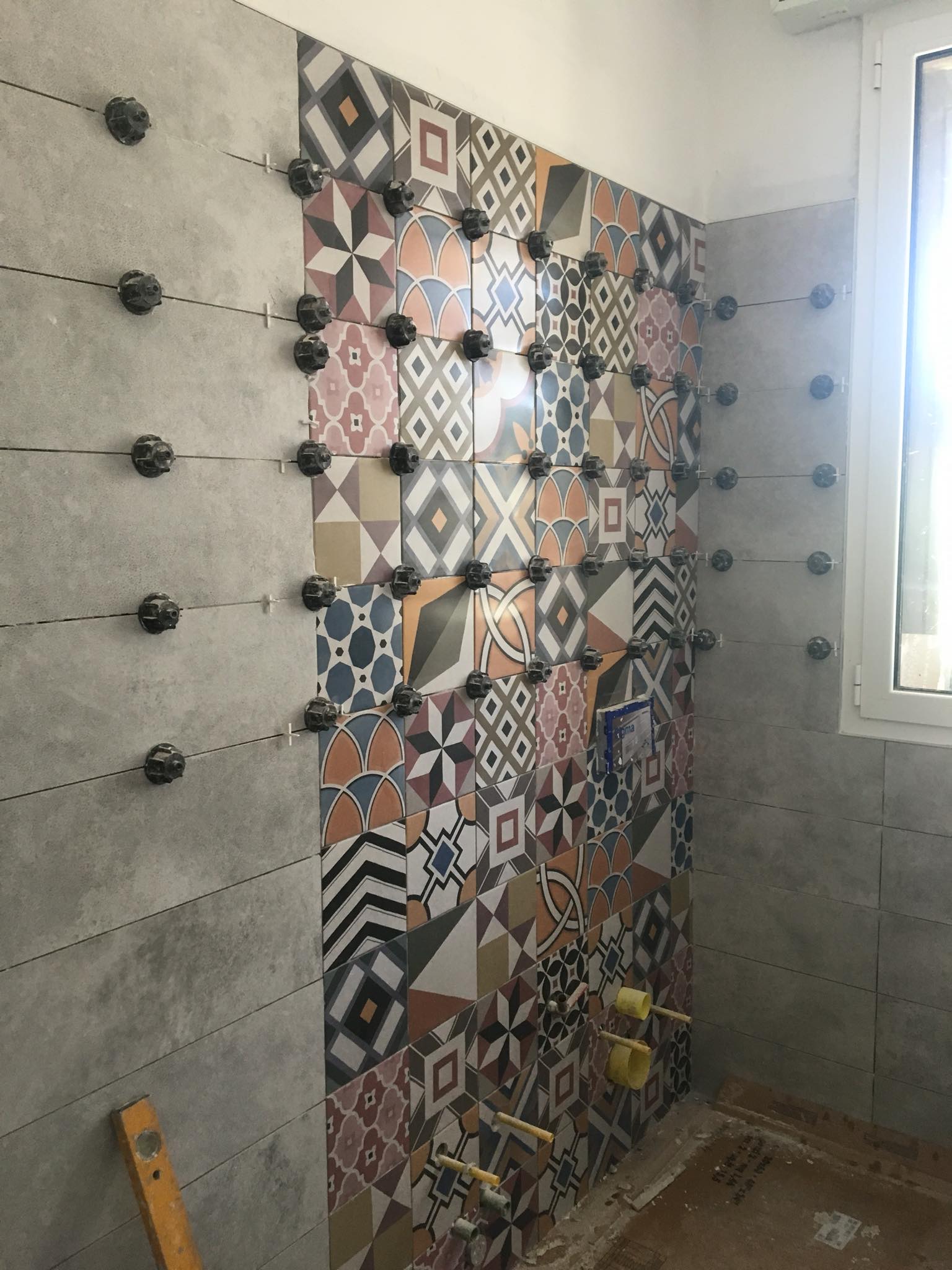 Parete del bagno parzialmente piastrellata con piastrelle grigie e a motivi, distanziatori per piastrelle già installati.