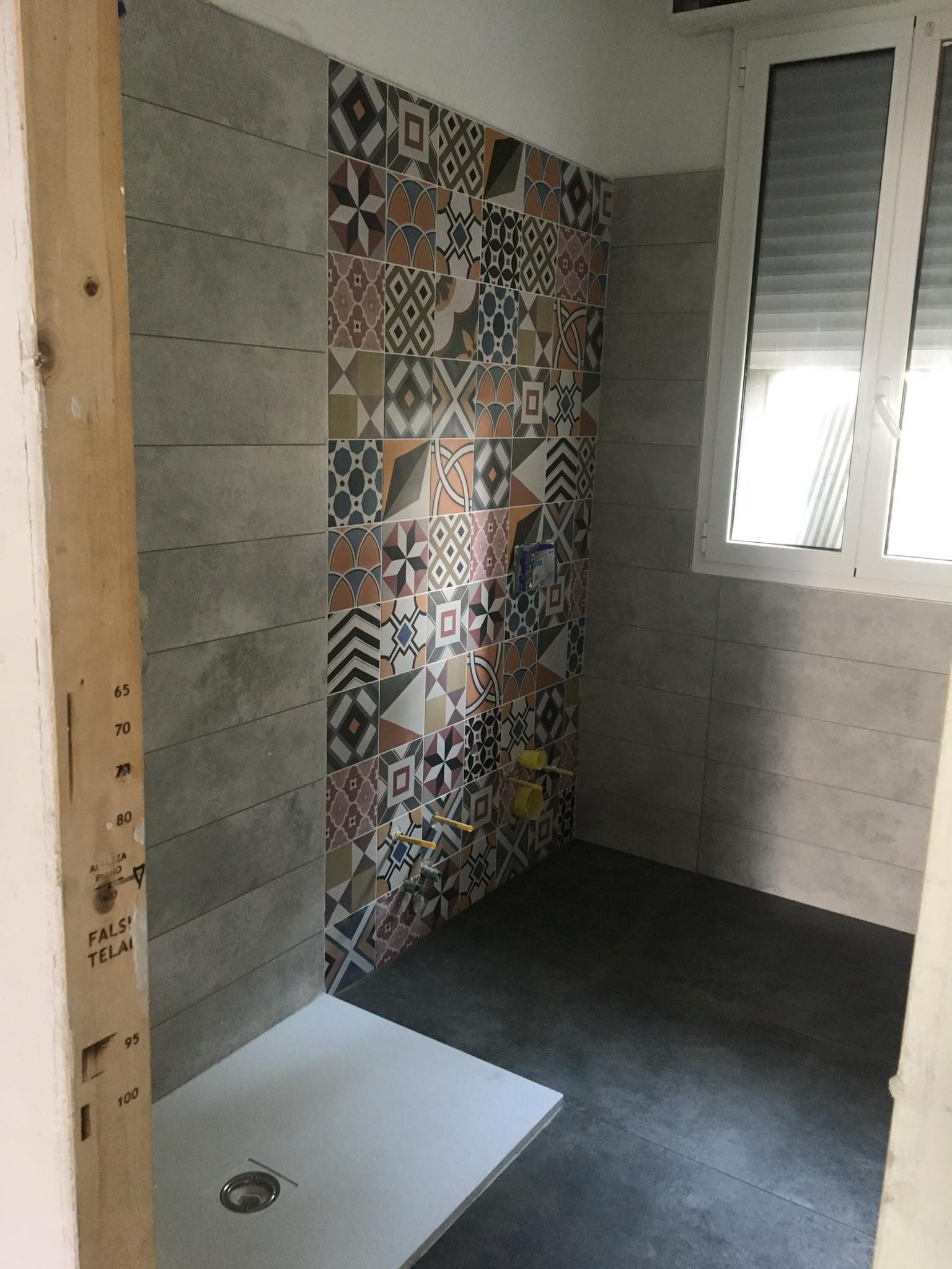 Bagno con pareti grigie, piastrelle decorative, piatto doccia, finestra con persiane e pavimento scuro.