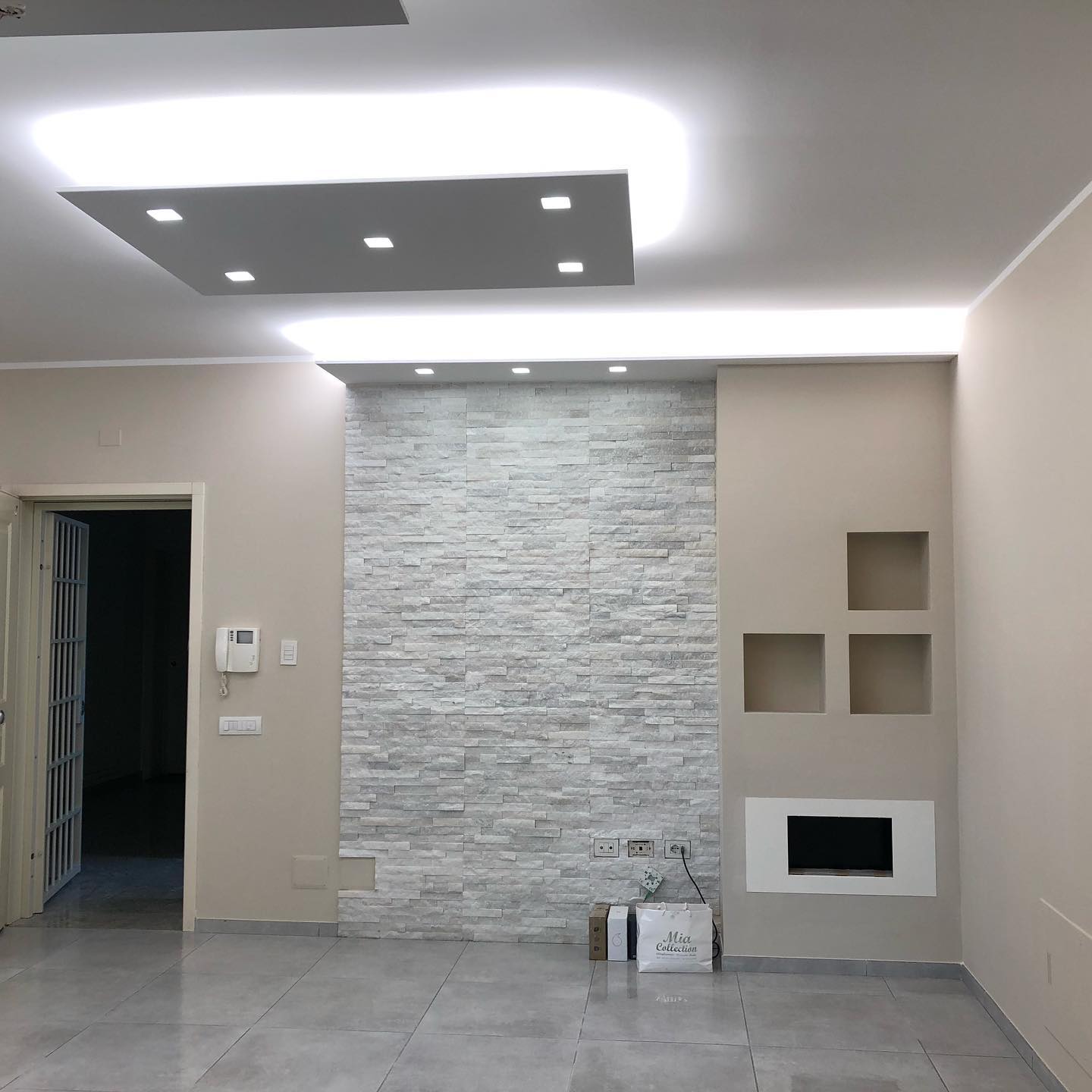 Soggiorno vuoto con parete bianca testurizzata, illuminazione da incasso e pavimento in piastrelle grigie.