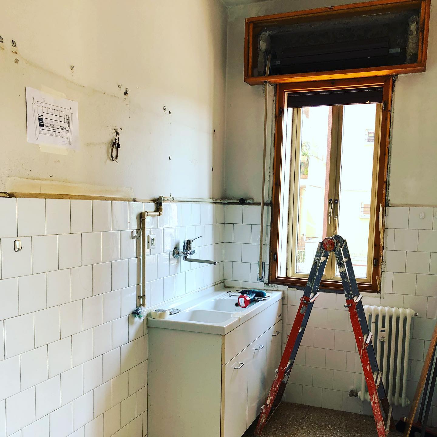 Cucina in fase di ristrutturazione con lavello, finestra, scala e pareti piastrellate.