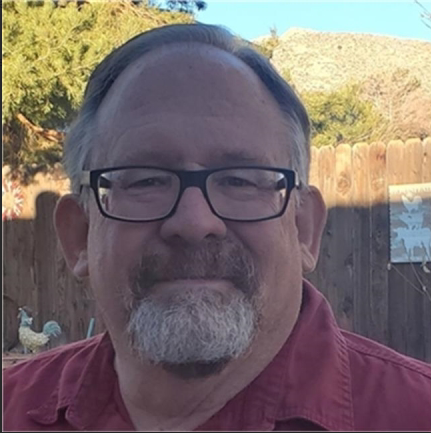 Man with beard, smiling, arms crossed, wearing tan shirt, standing outside.