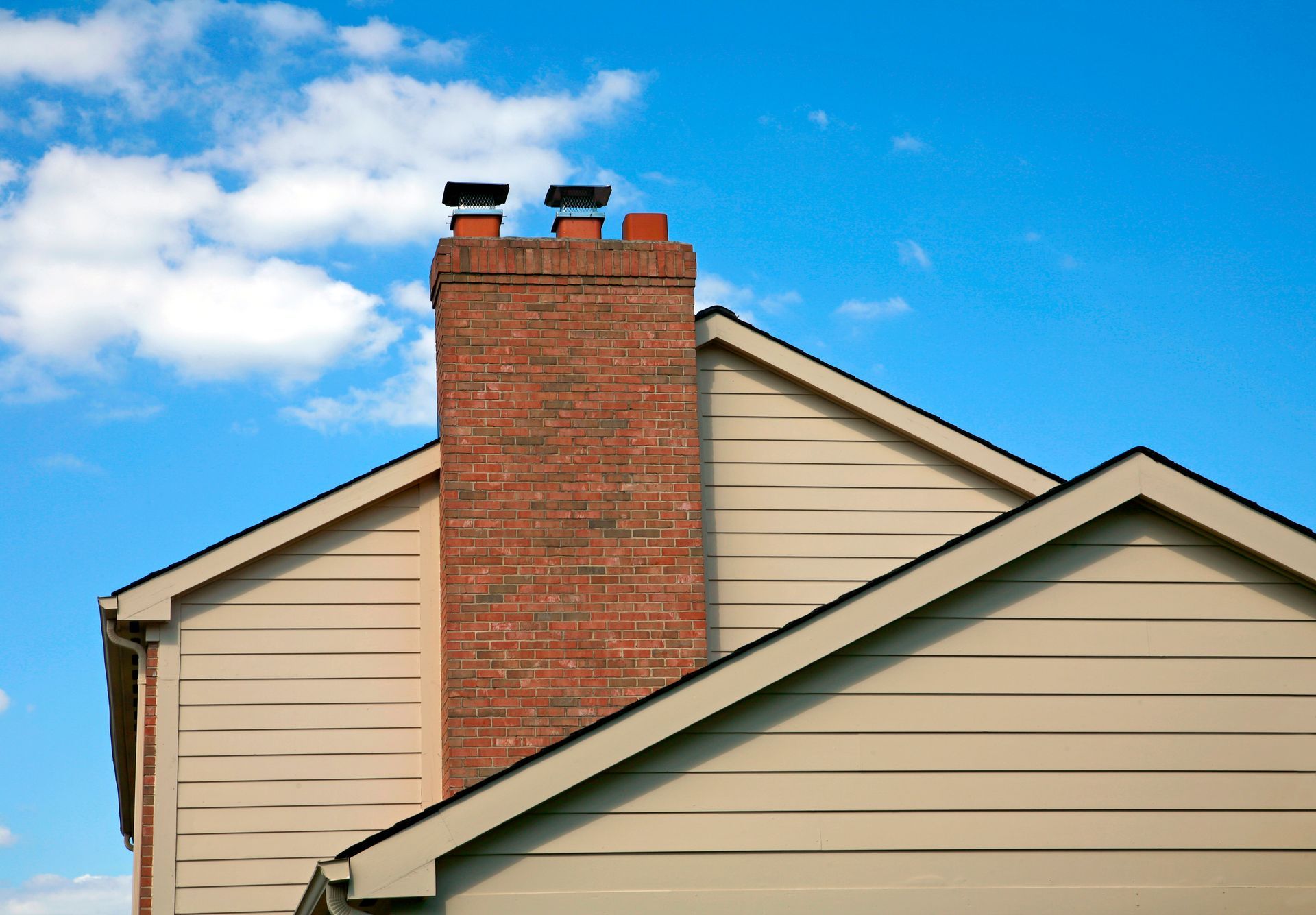 Brick chimney rising from a tan house roof against a blue sky with clouds.