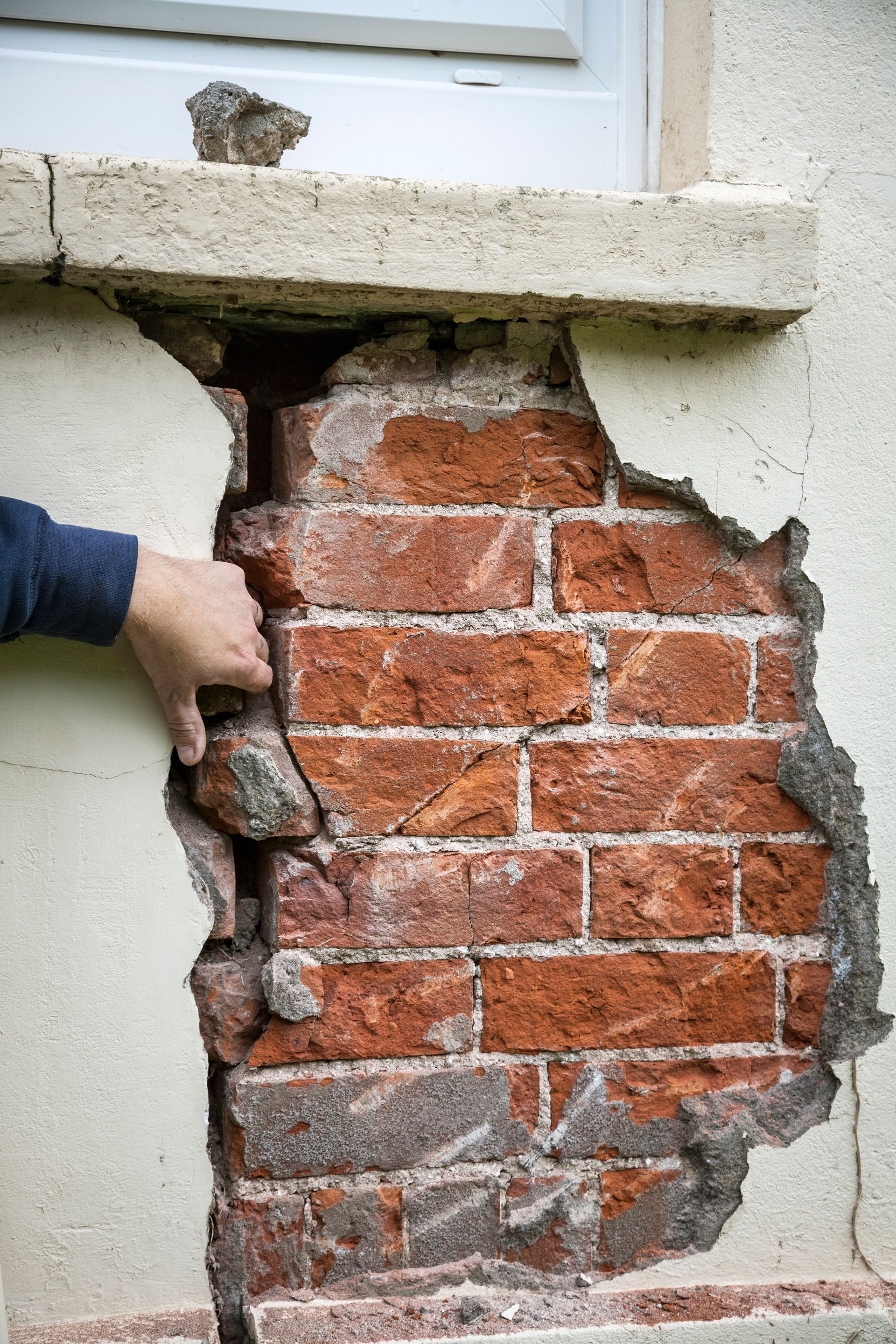 Damaged exterior wall exposing red bricks. A hand touches the crumbling plaster near a window.