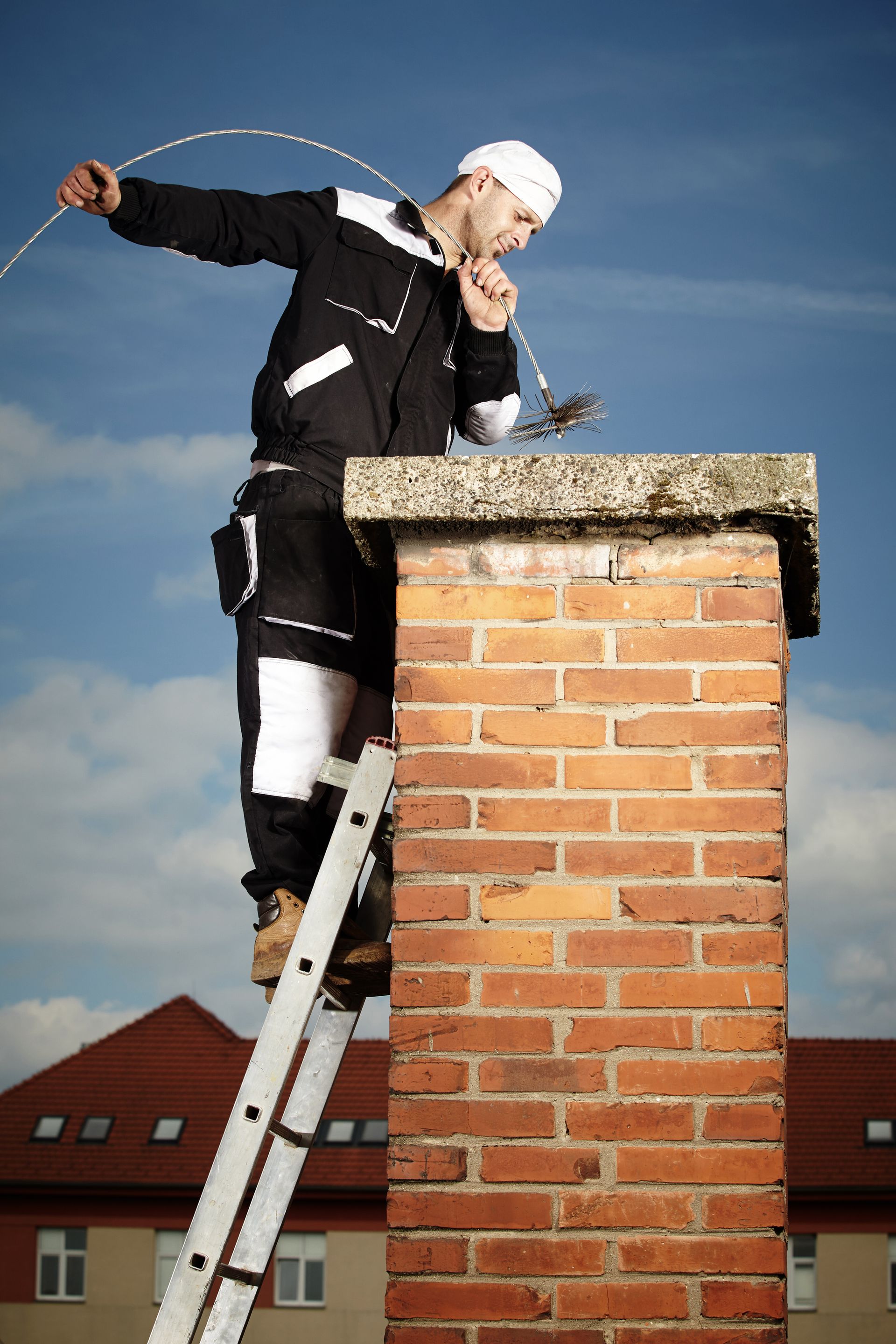 Chimney sweep on a roof using a brush to clean a chimney against a blue sky.