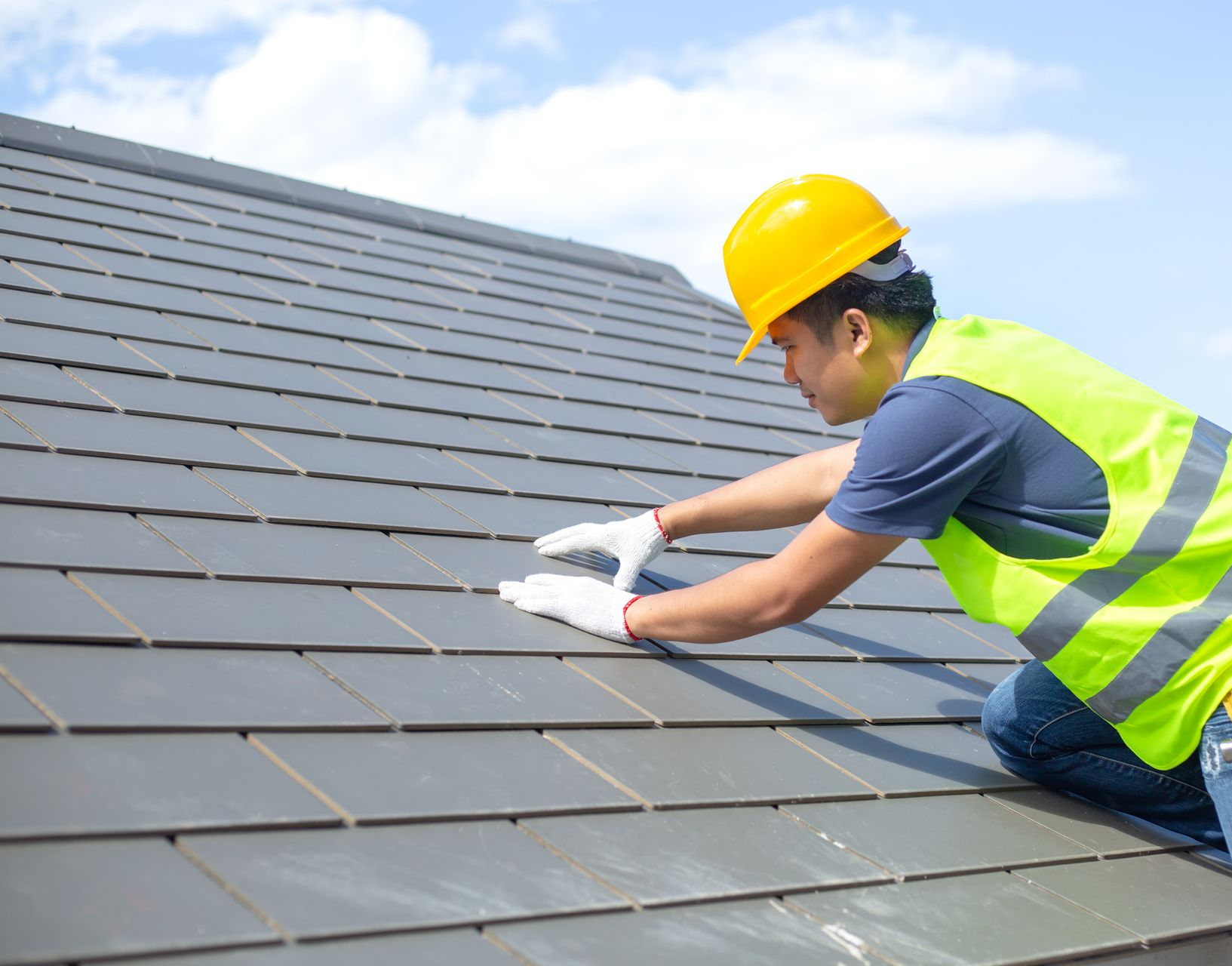 Roofer in safety gear inspecting a gray tiled roof under a blue sky.