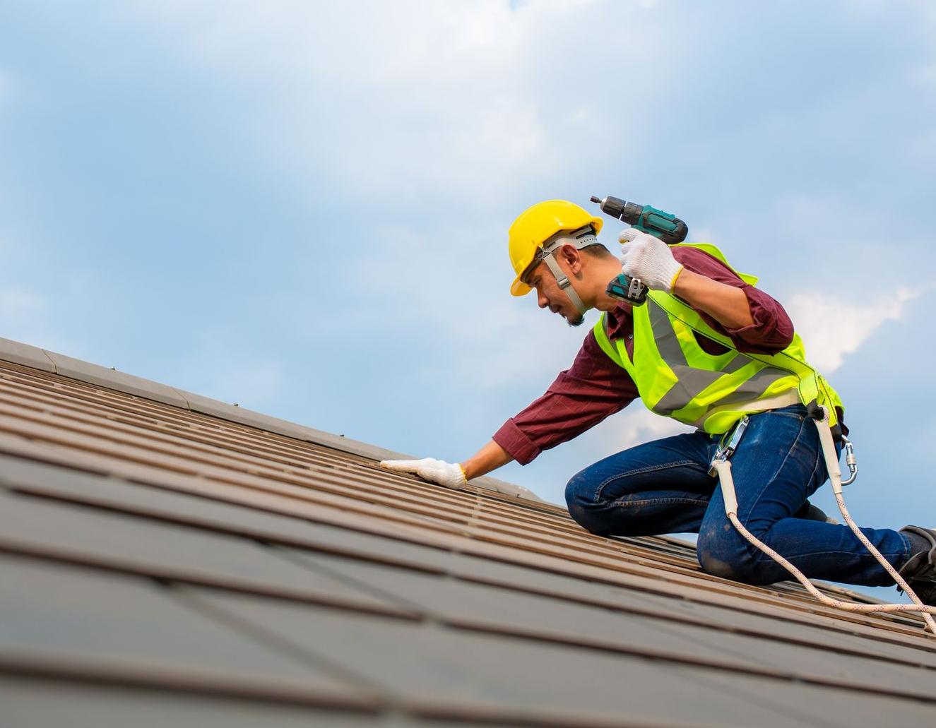 Roofer on a sloped roof wearing a hard hat, safety vest, and harness, using a drill on shingles under a cloudy sky.