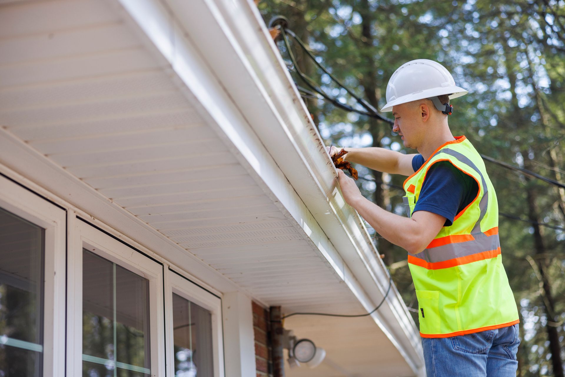 Person in safety vest and hard hat working on a roof gutter near windows.
