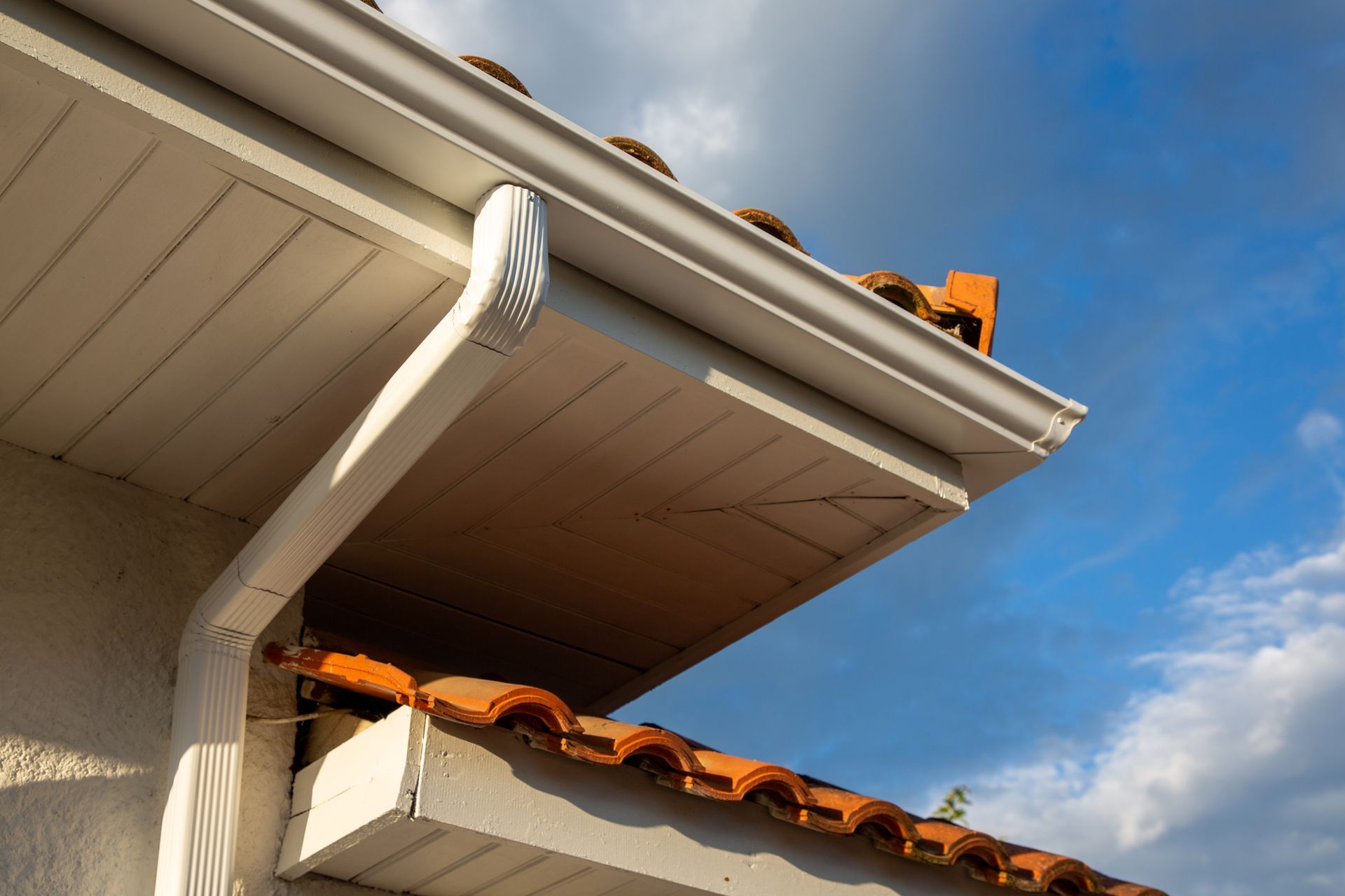 White gutters attached to a white soffit on a building with terracotta roof tiles against a cloudy sky.