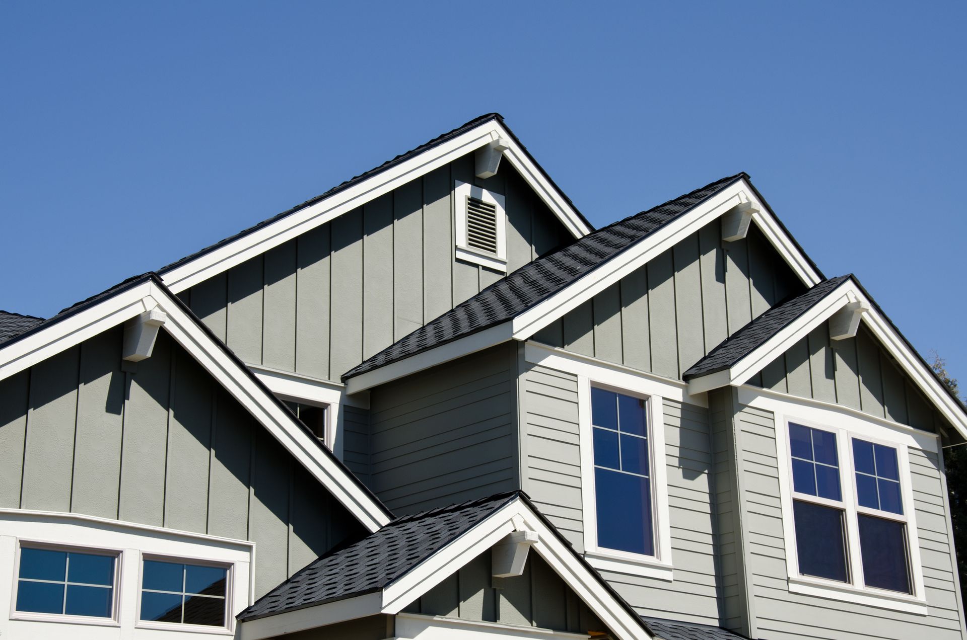 Gray house with multiple rooflines, white trim, and blue windows against a clear sky.