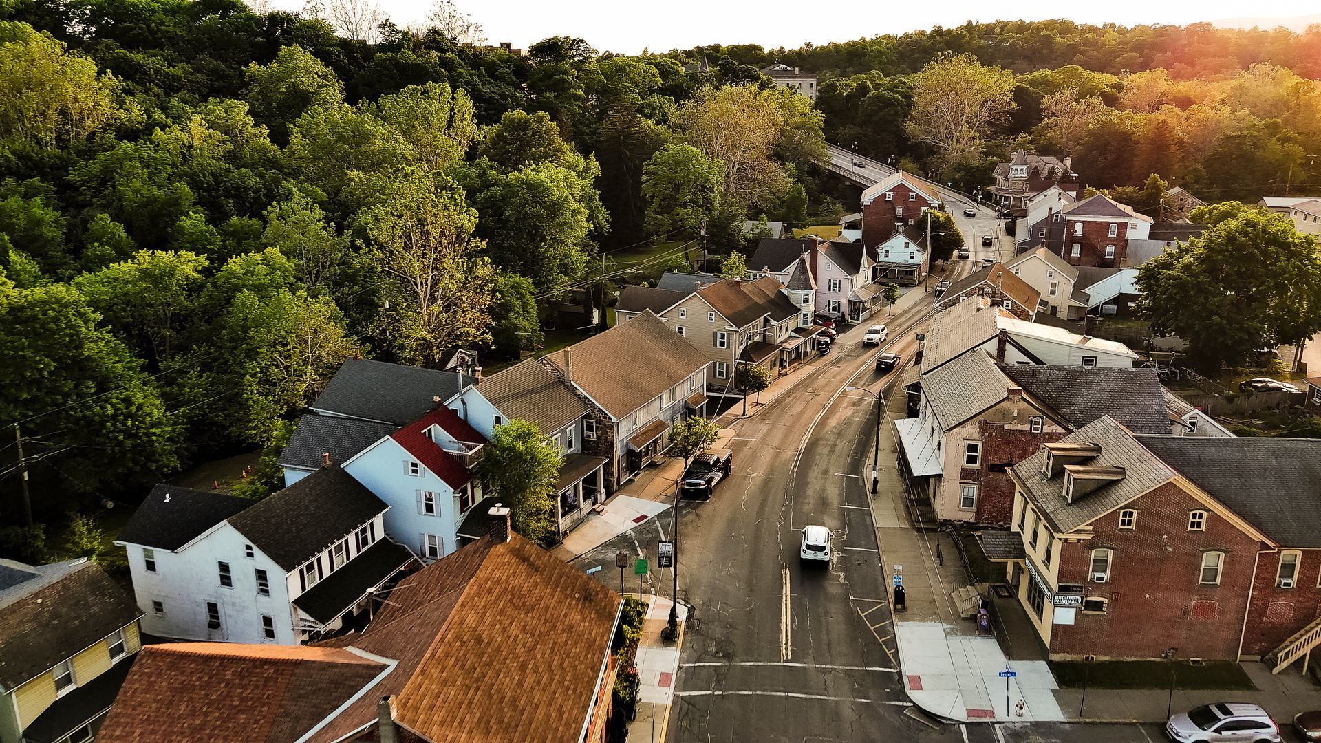 Aerial view of a quaint town with a road, buildings, and lush green trees. Sunlight bathes the scene.