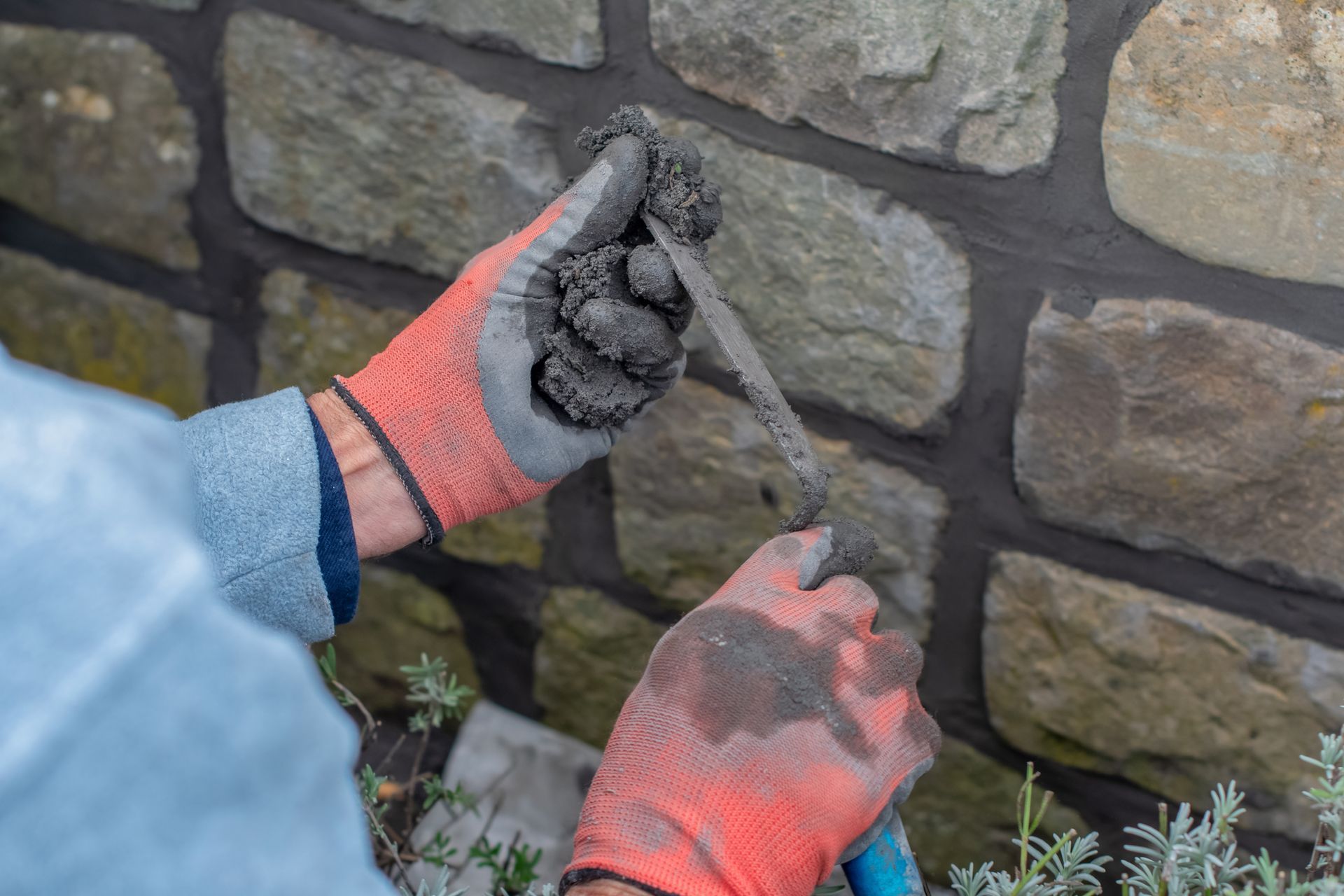 Hands in orange gloves using a trowel to apply dark mortar between stone wall blocks.