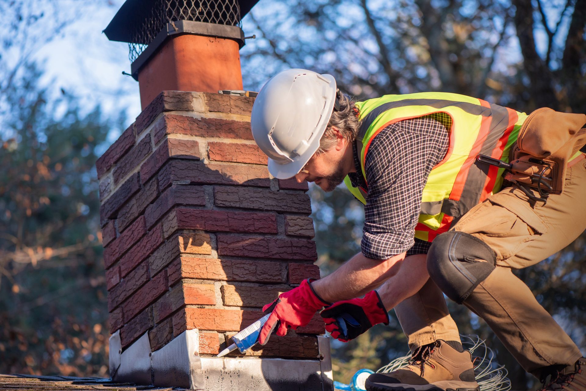 Construction worker repairing brick chimney on a roof, using sealant. Wearing safety gear.
