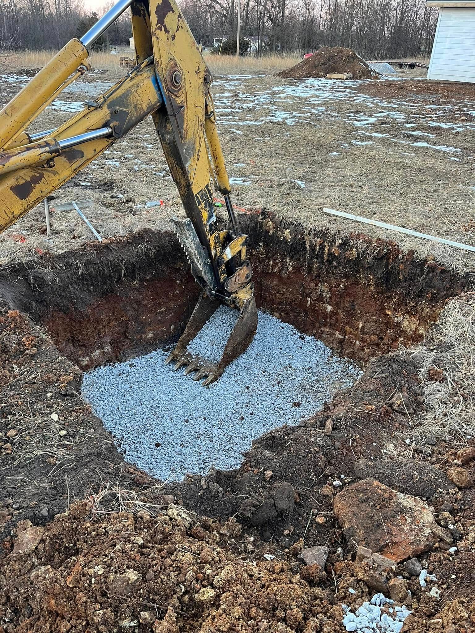 Yellow backhoe bucket dumping gravel into a square excavation pit.