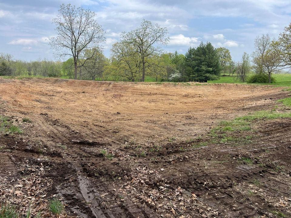Dirt-filled basin, recently excavated, in a grassy field, trees in the background, overcast sky.