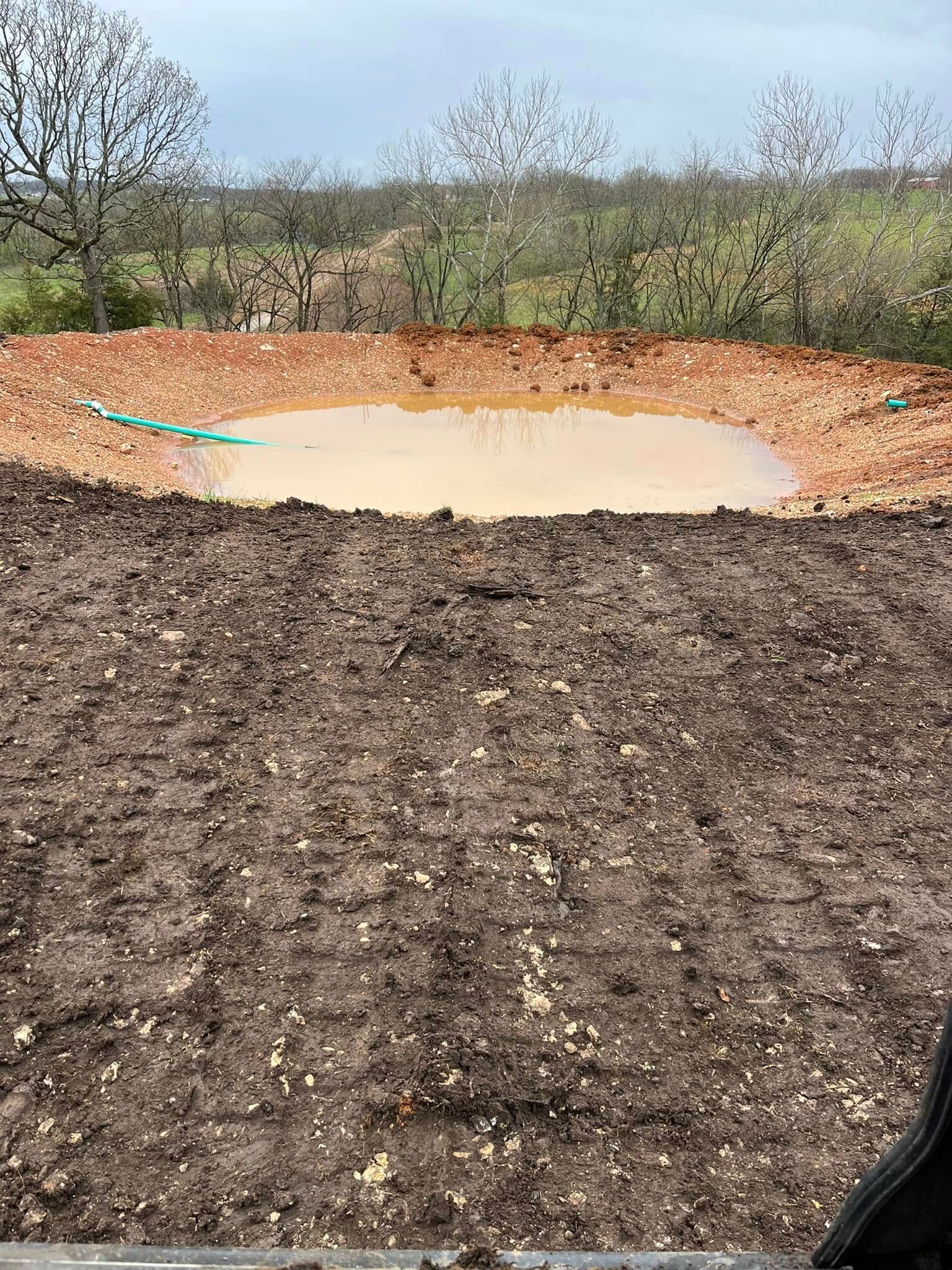 Earthen pond with muddy water, surrounded by red soil and dirt field; trees in the background.