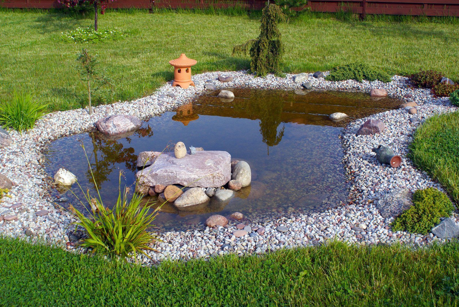 A decorative backyard pond surrounded by gravel, rocks, and green grass.