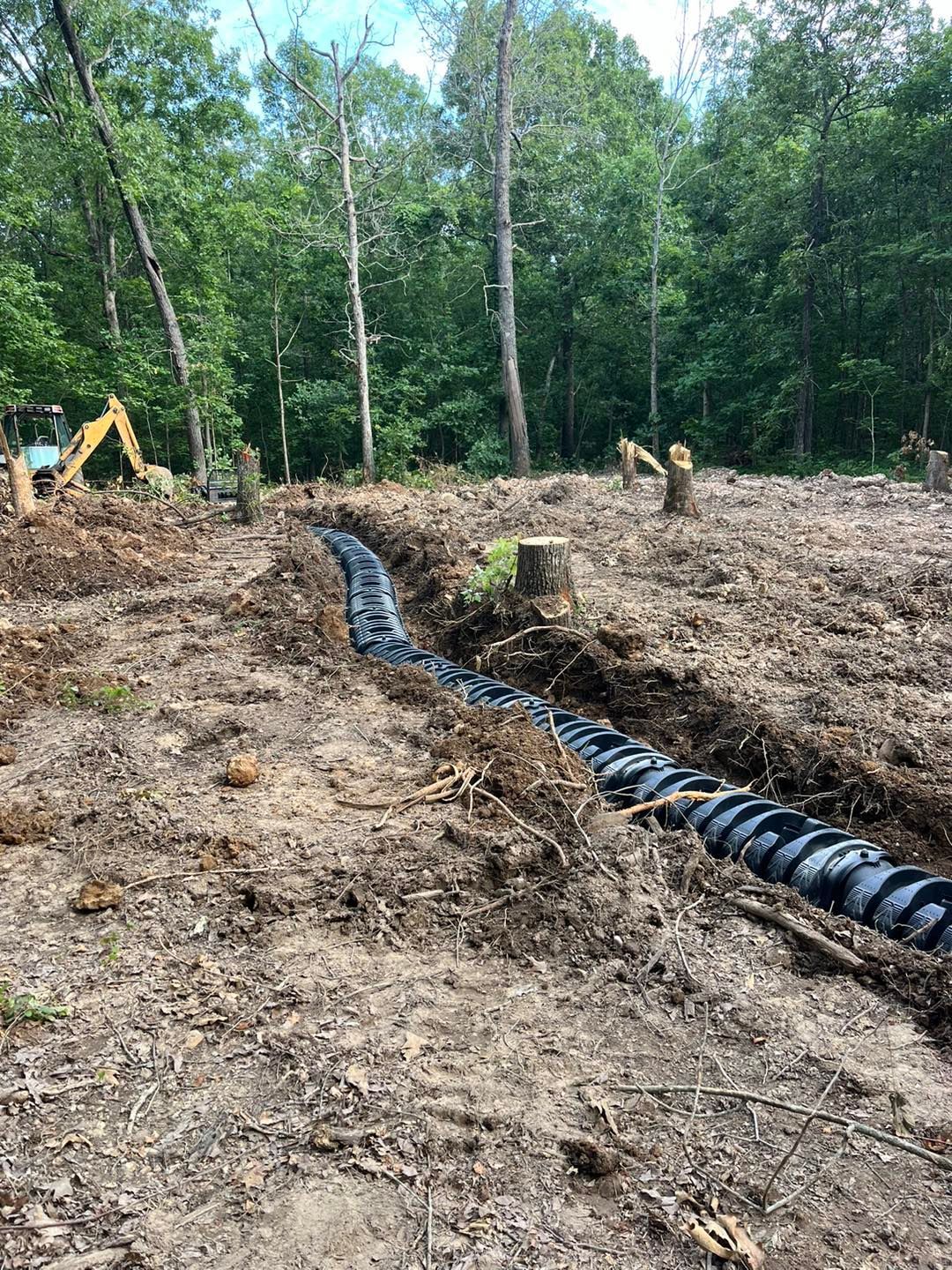 Black pipe in a trench, trees in background; possible septic system installation in a wooded area.