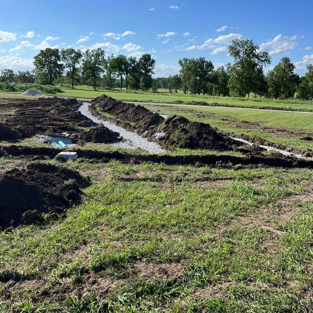 Trenches dug in a grassy field for construction, with piles of dirt and trees in the background under a blue sky.