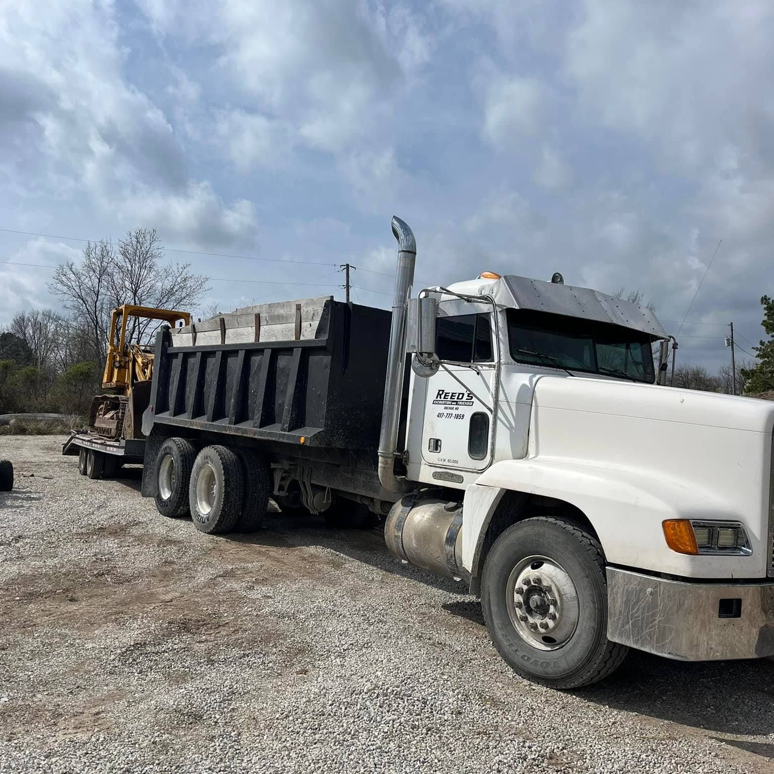 White dump truck hauling a trailer with construction equipment on a gravel lot under a cloudy sky.