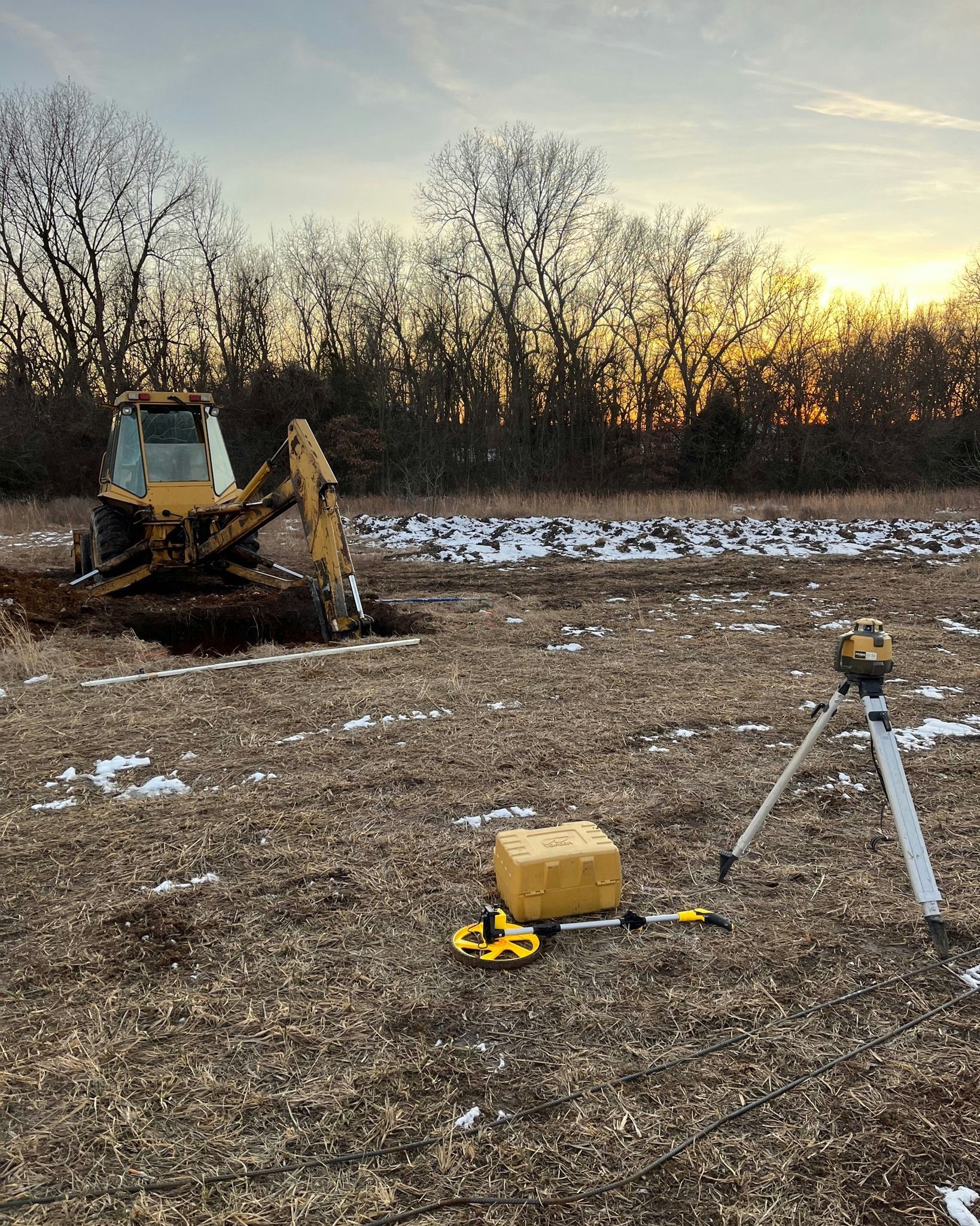 Backhoe digging in a field at sunset, with survey equipment nearby.