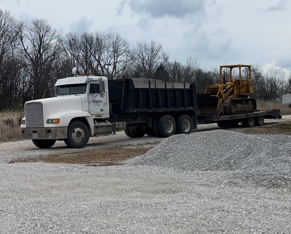 White dump truck hauling a black bed filled with gravel and a yellow bulldozer on a trailer, parked on gravel near a pile of gravel.