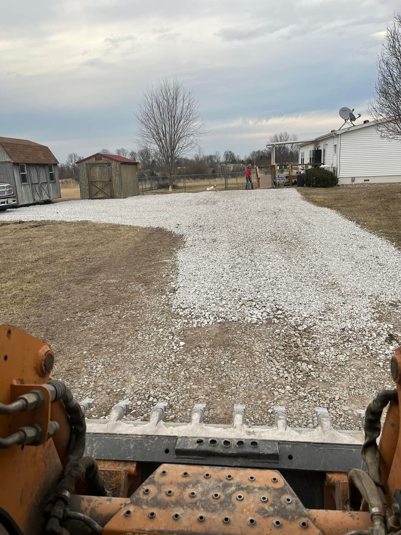 View from a skid steer, showing a gravel area being constructed near a house and outbuildings.