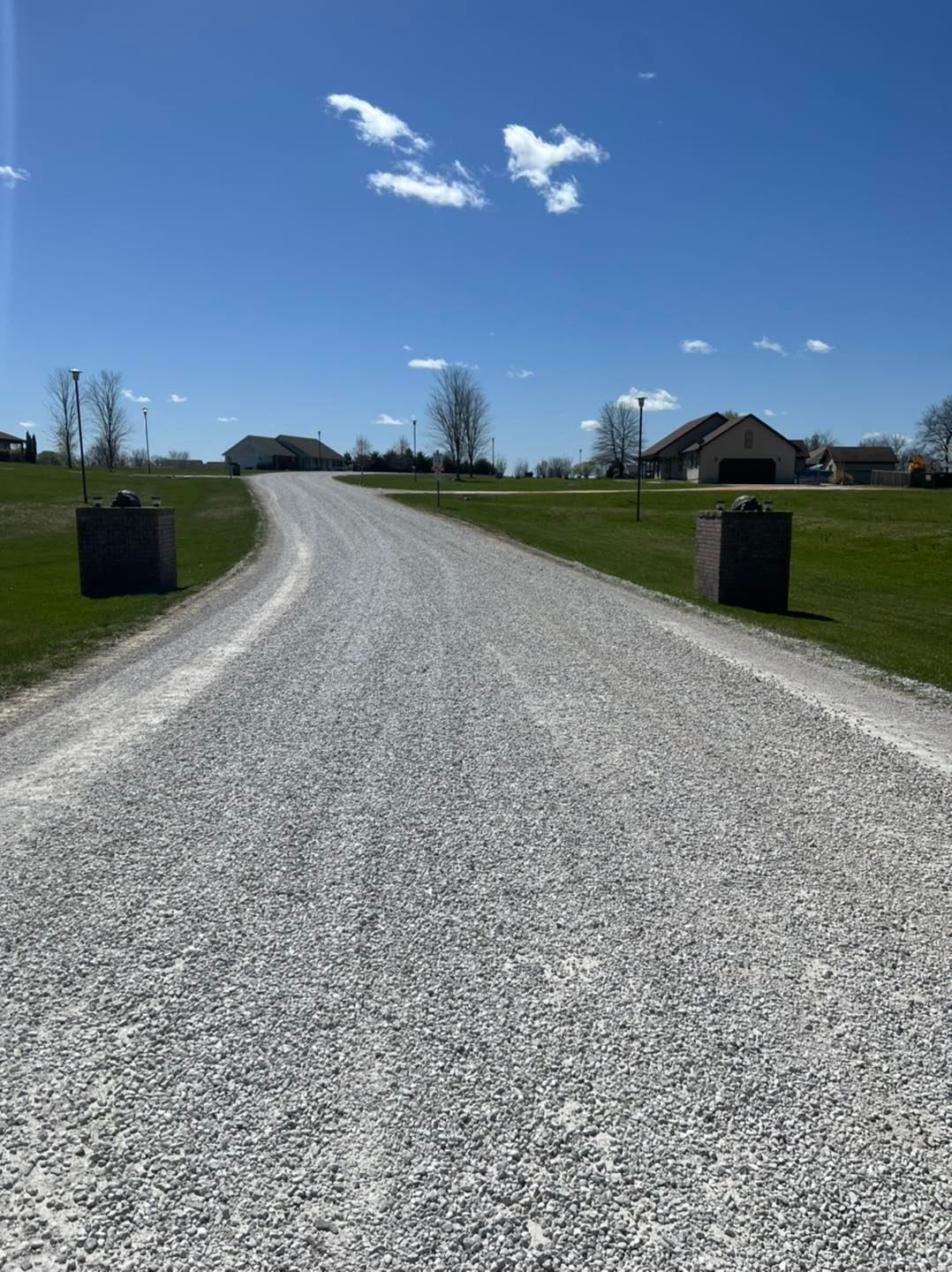 Gravel driveway curving uphill toward houses on a sunny day. Green grass borders the drive.