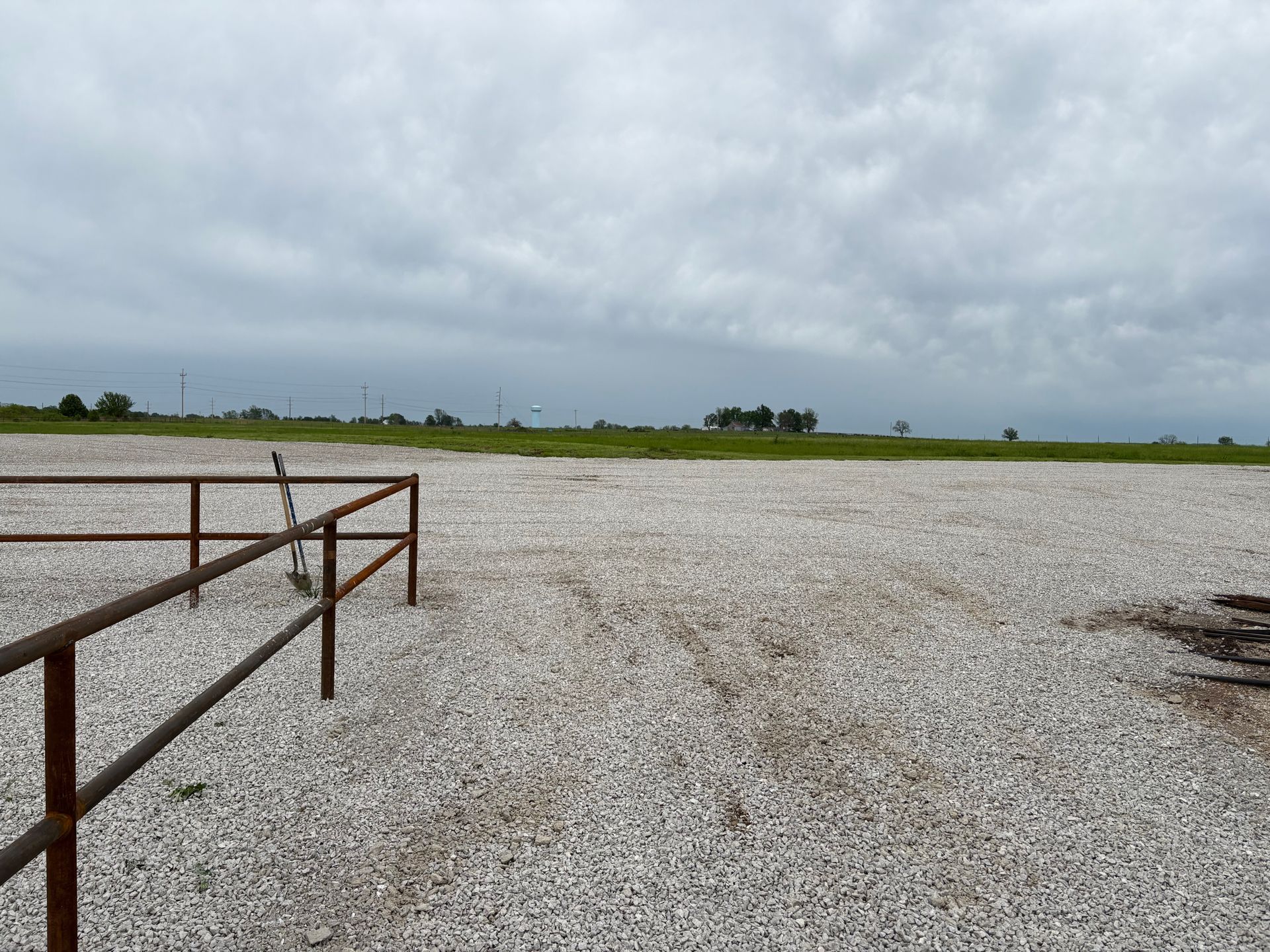 Gravel lot with rusty metal fence, green field, and cloudy sky.