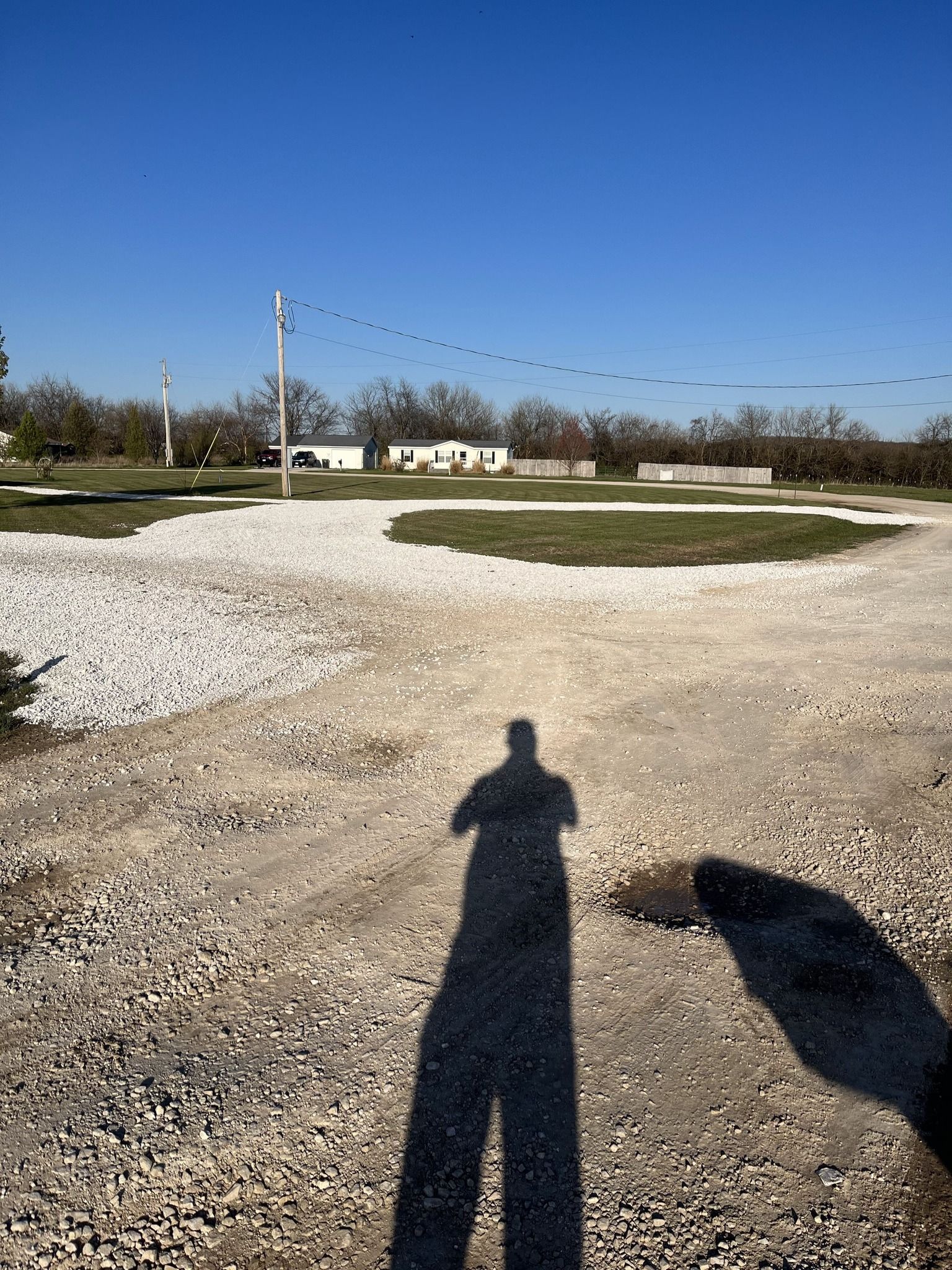 Person's shadow on a gravel path, with green areas and a white building in the background on a sunny day.