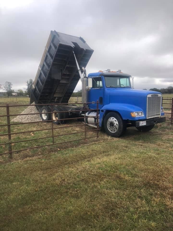 Blue dump truck raised to release gravel onto a grassy field.