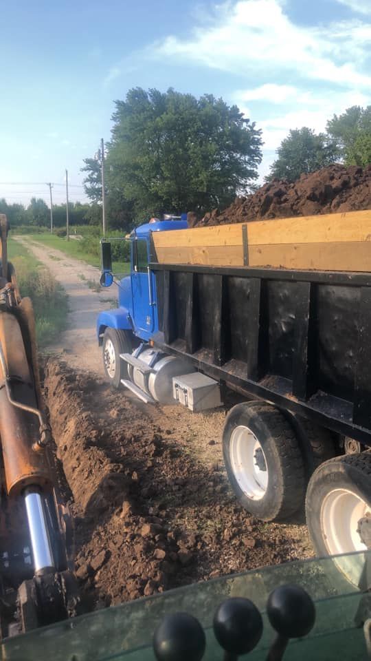 Blue dump truck dumping soil into a trench beside a dirt road. Green tractor controls are in the foreground.
