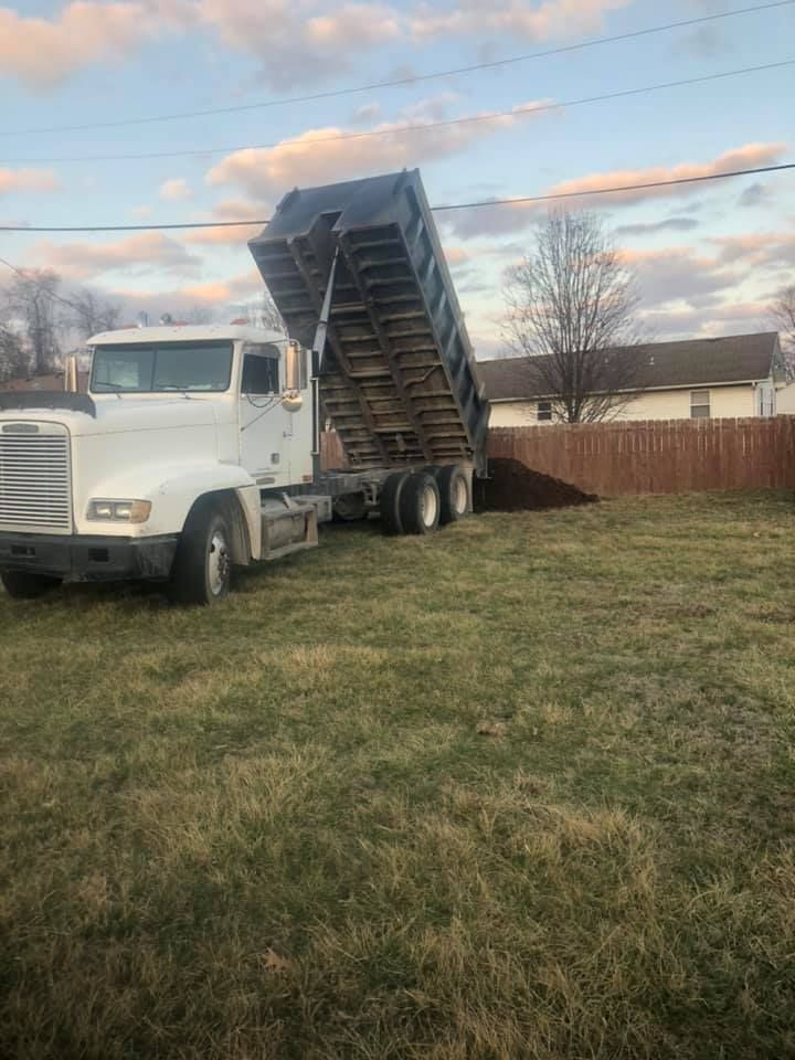 A white dump truck in a grassy yard is raising its bed to dump dark soil near a fence under a cloudy sky.
