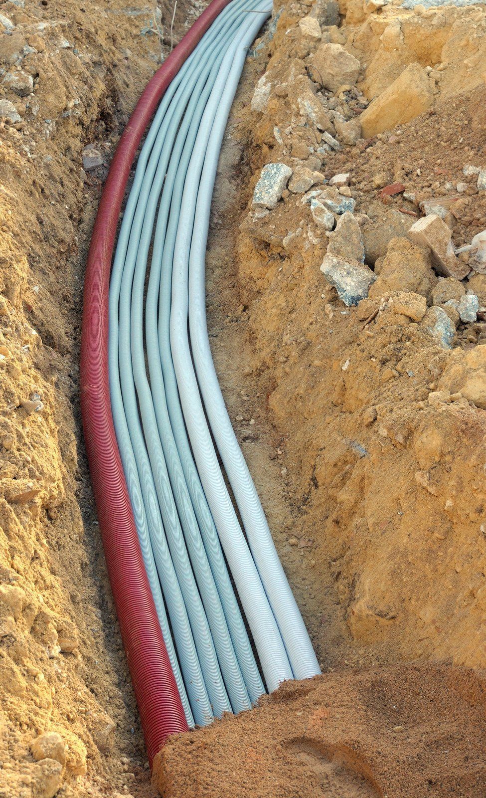 Trench with multiple gray and one red corrugated conduit pipes buried in the soil.