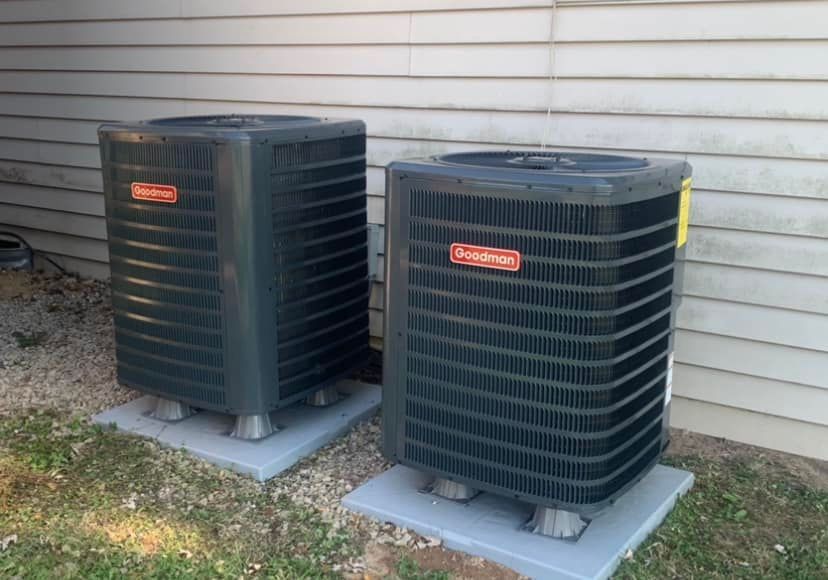 Two Goodman air conditioning units on concrete pads beside a building.
