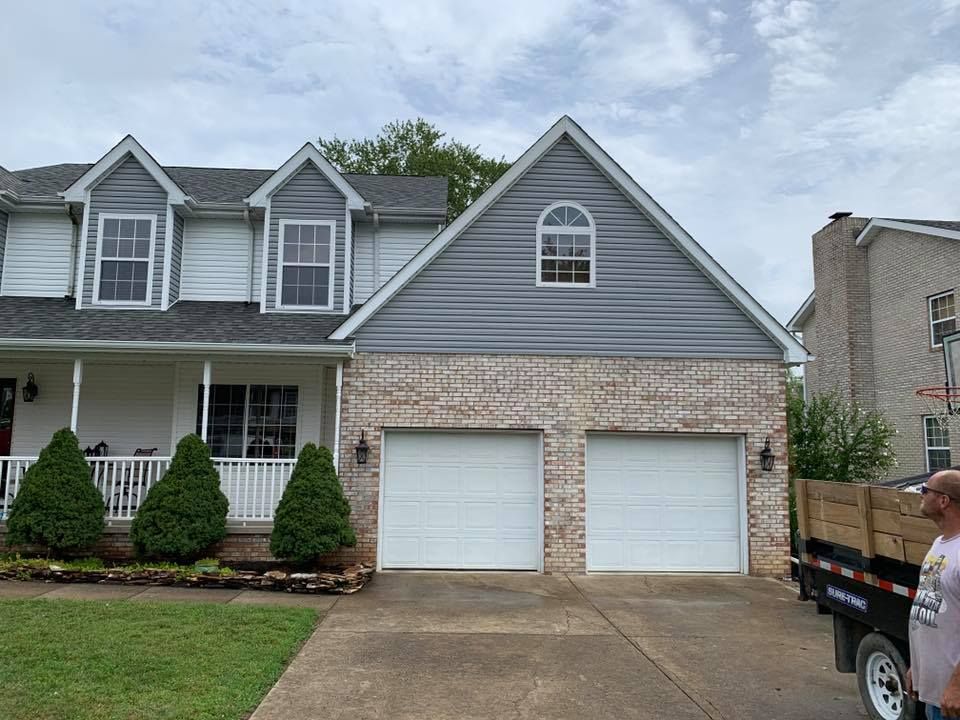 Two-car garage with brick base and gray siding above, connected to a white house with dormers; person on the right.