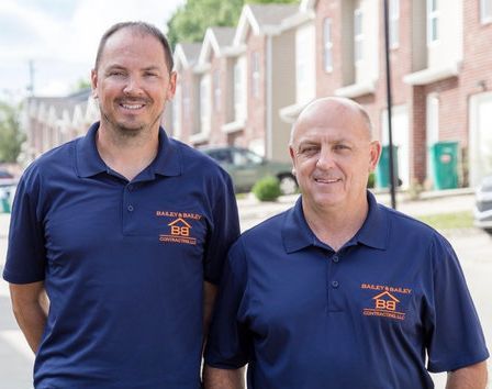 Two men in navy polo shirts with a logo, smiling in front of townhomes on a sunny day.