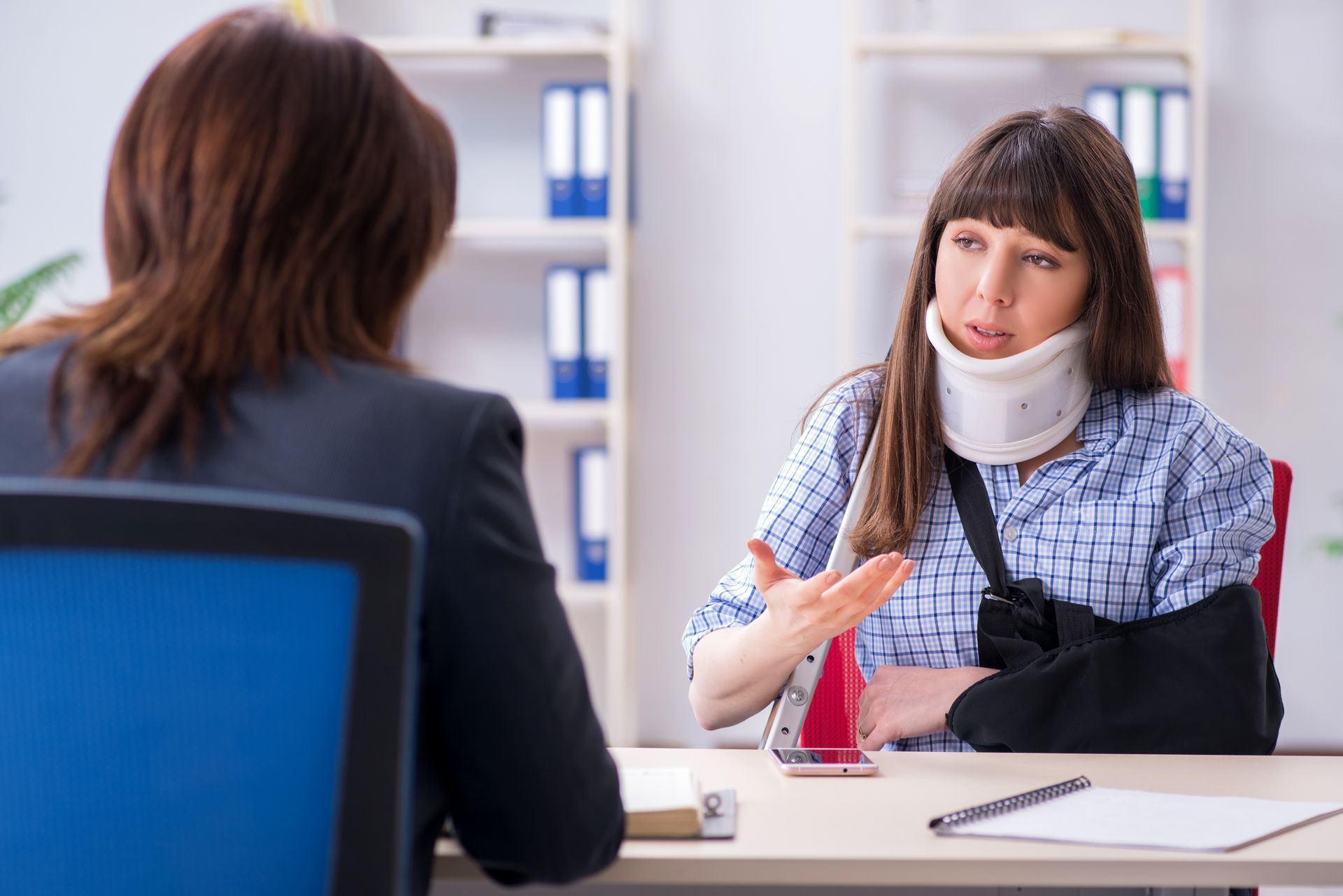 Woman with arm sling and neck brace talking to a person at a desk in an office setting.