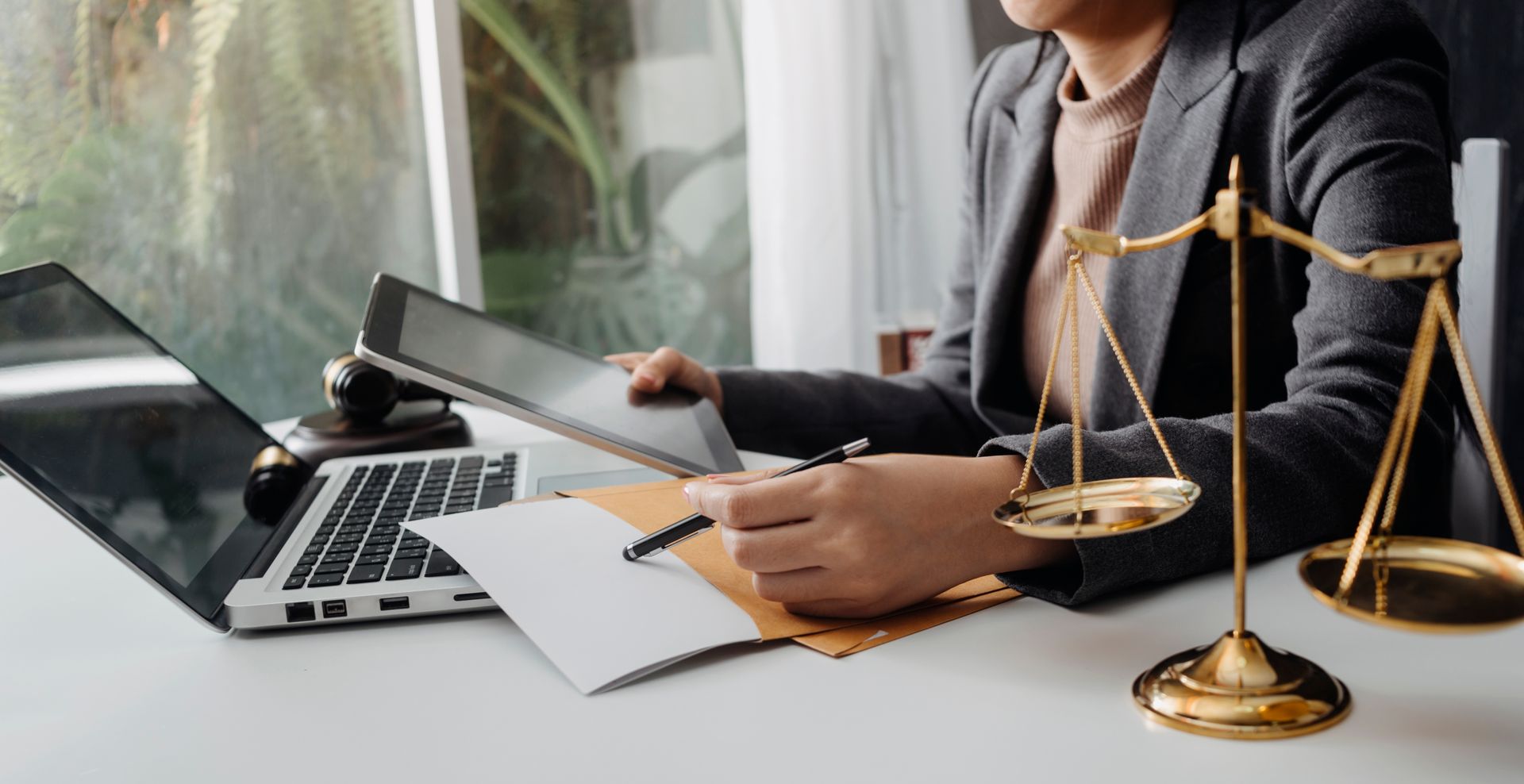 Woman in suit working at desk with laptop, tablet, law scale, and documents.