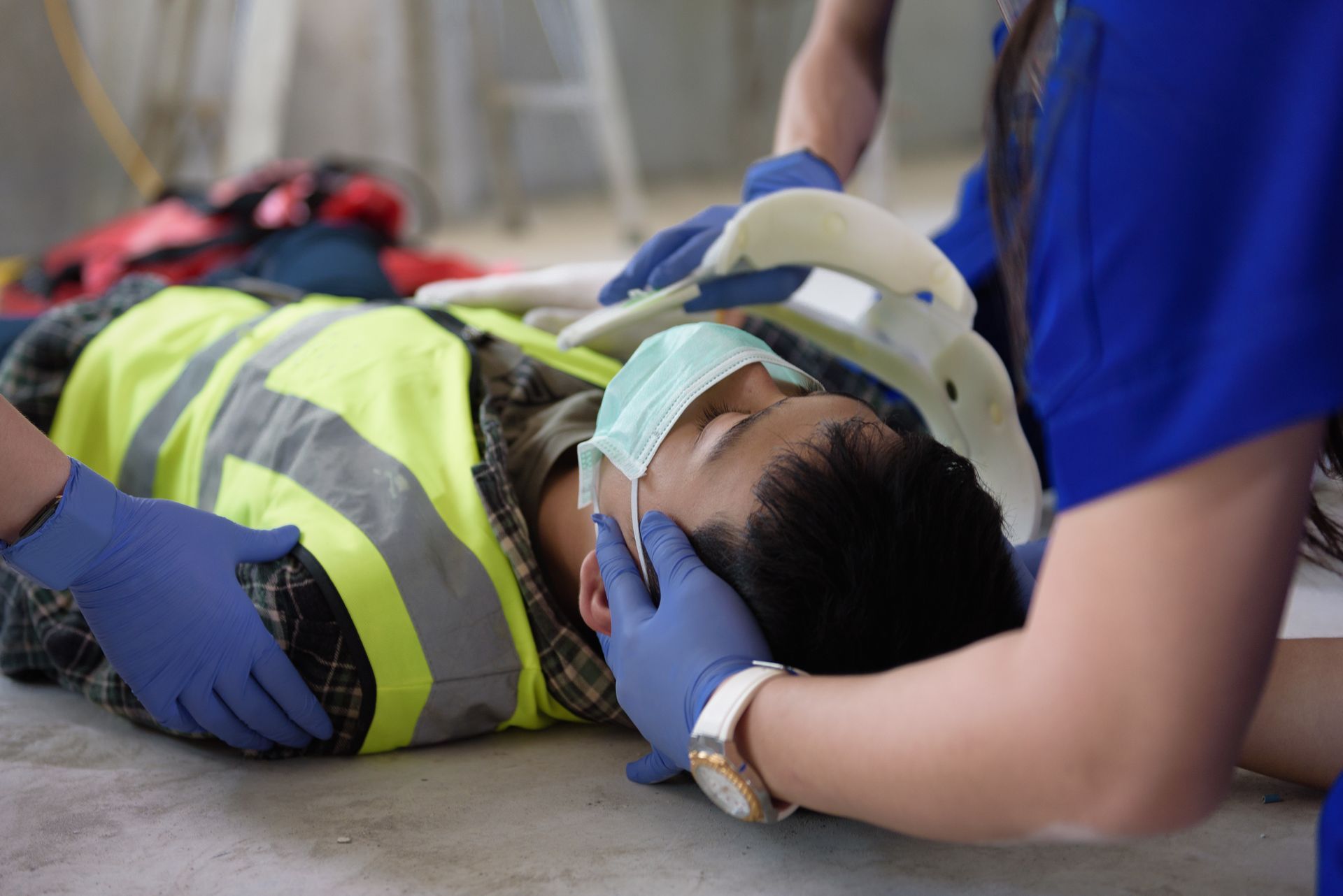 Two people tending to a person on the ground, wearing a neck brace, mask, and construction vest.