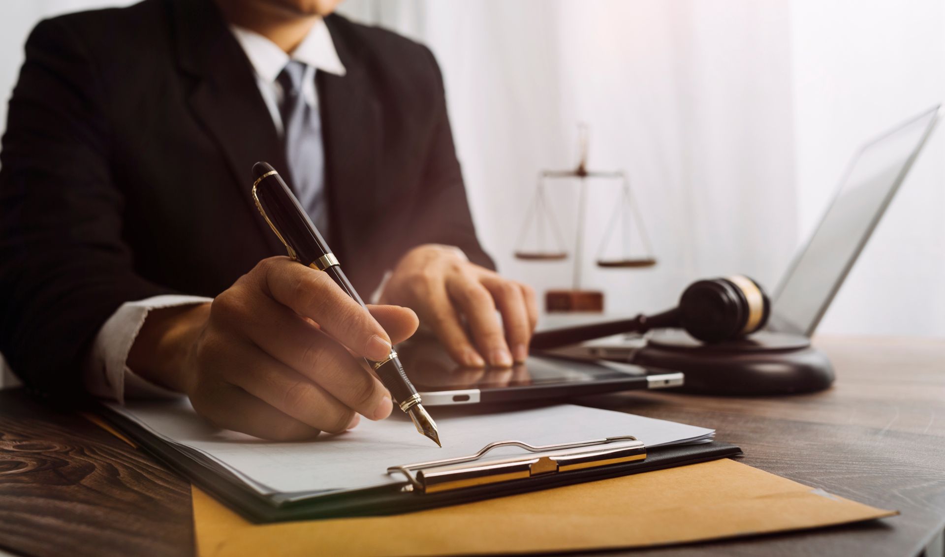 Person in suit writing on a notepad, with scales of justice, gavel, and laptop on a desk.