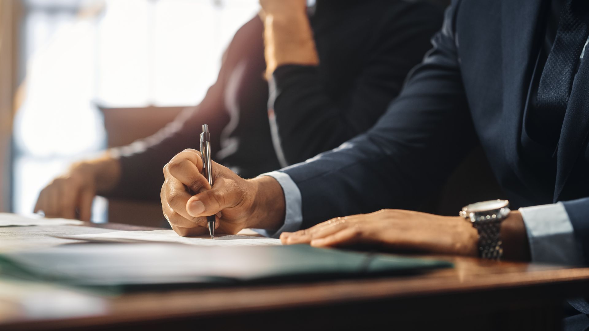 Person in suit writing at a table with another person, watch visible.