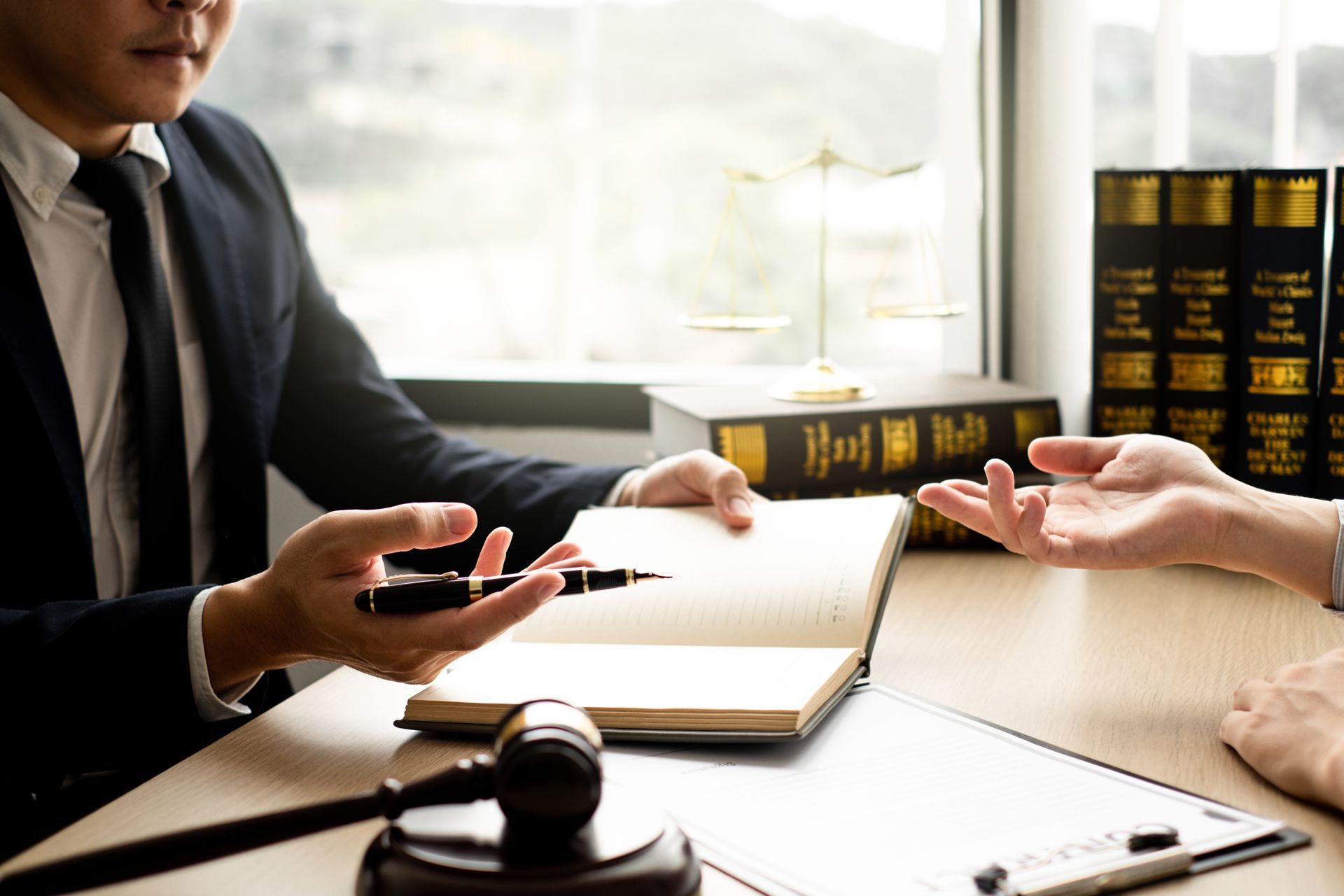 Lawyer in suit talking to client, open notebook, gavel, and books on desk.