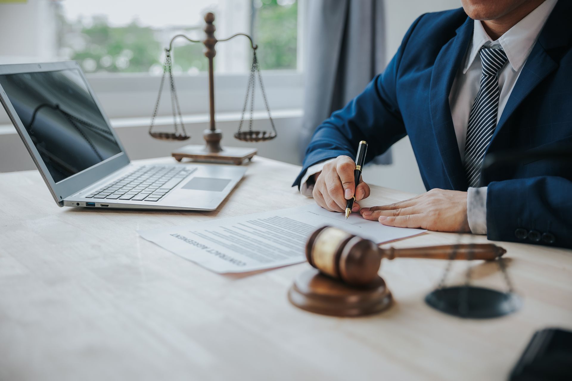 Lawyer signing documents, desk with laptop, gavel, and scales of justice.