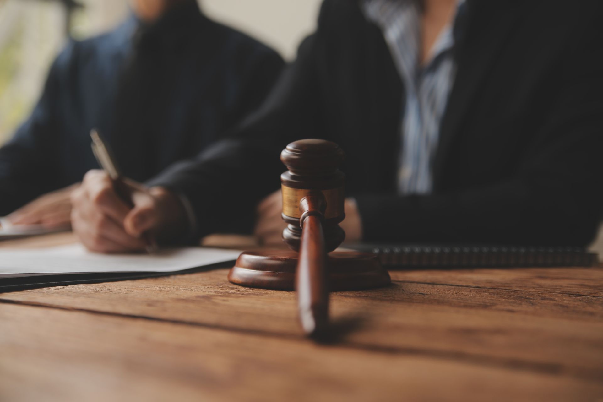 Gavel on a wooden table with two people in suits, one writing, in a legal setting.