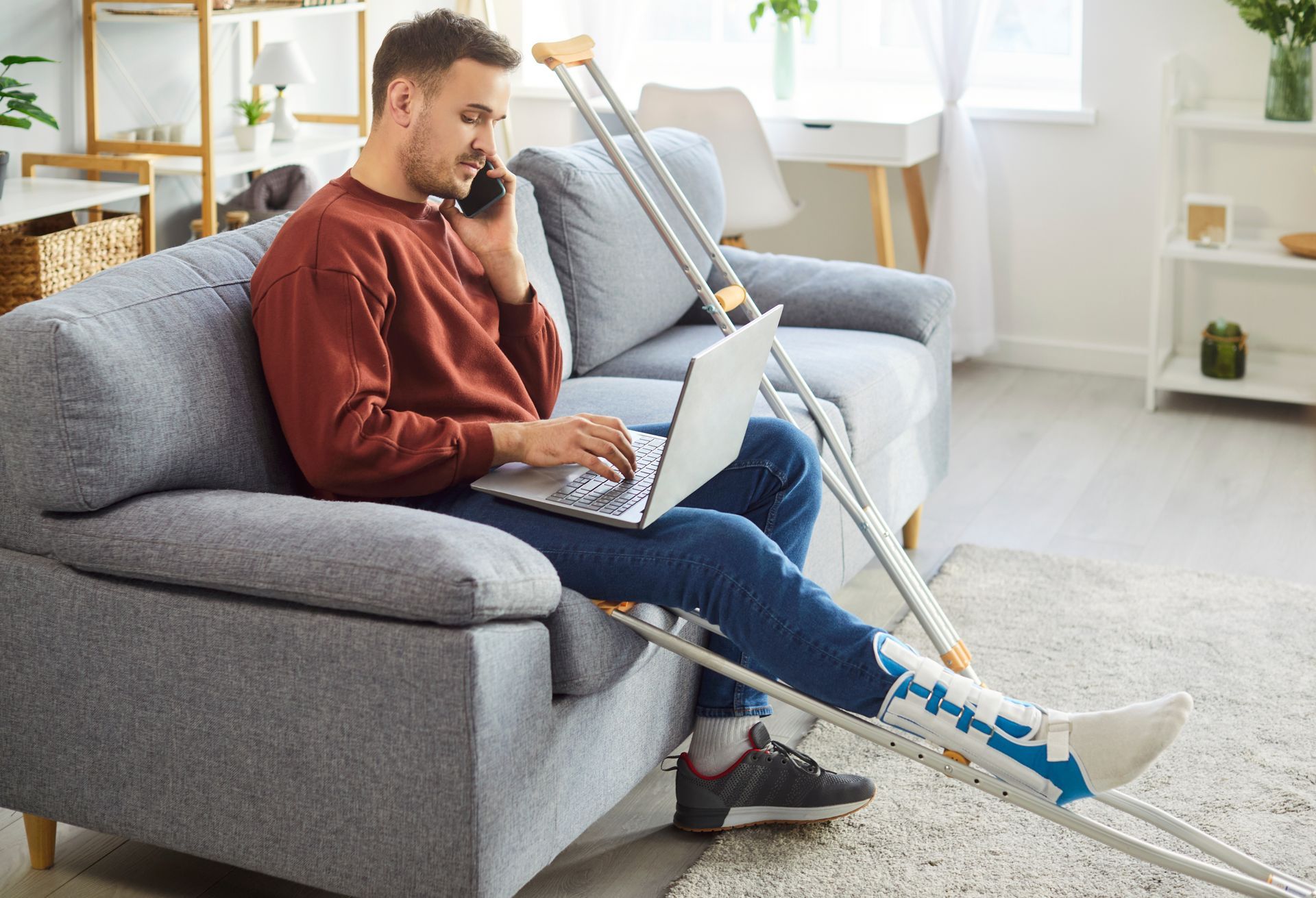 Man on couch with leg cast, working on laptop and talking on the phone. 