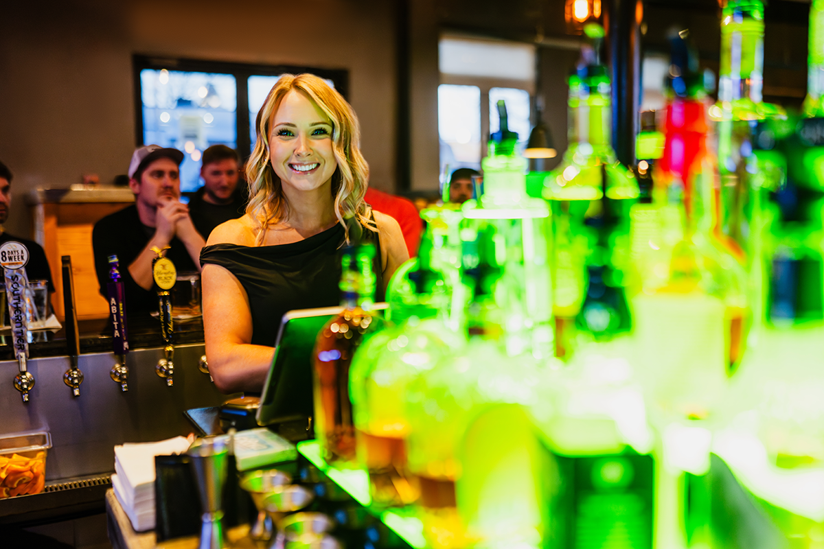 Bartender smiles behind a bar with illuminated liquor bottles; other people in background.