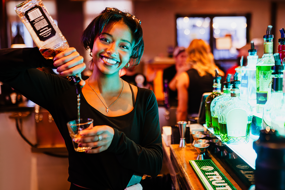 Bartender pouring a drink, smiling, behind a well-lit bar.