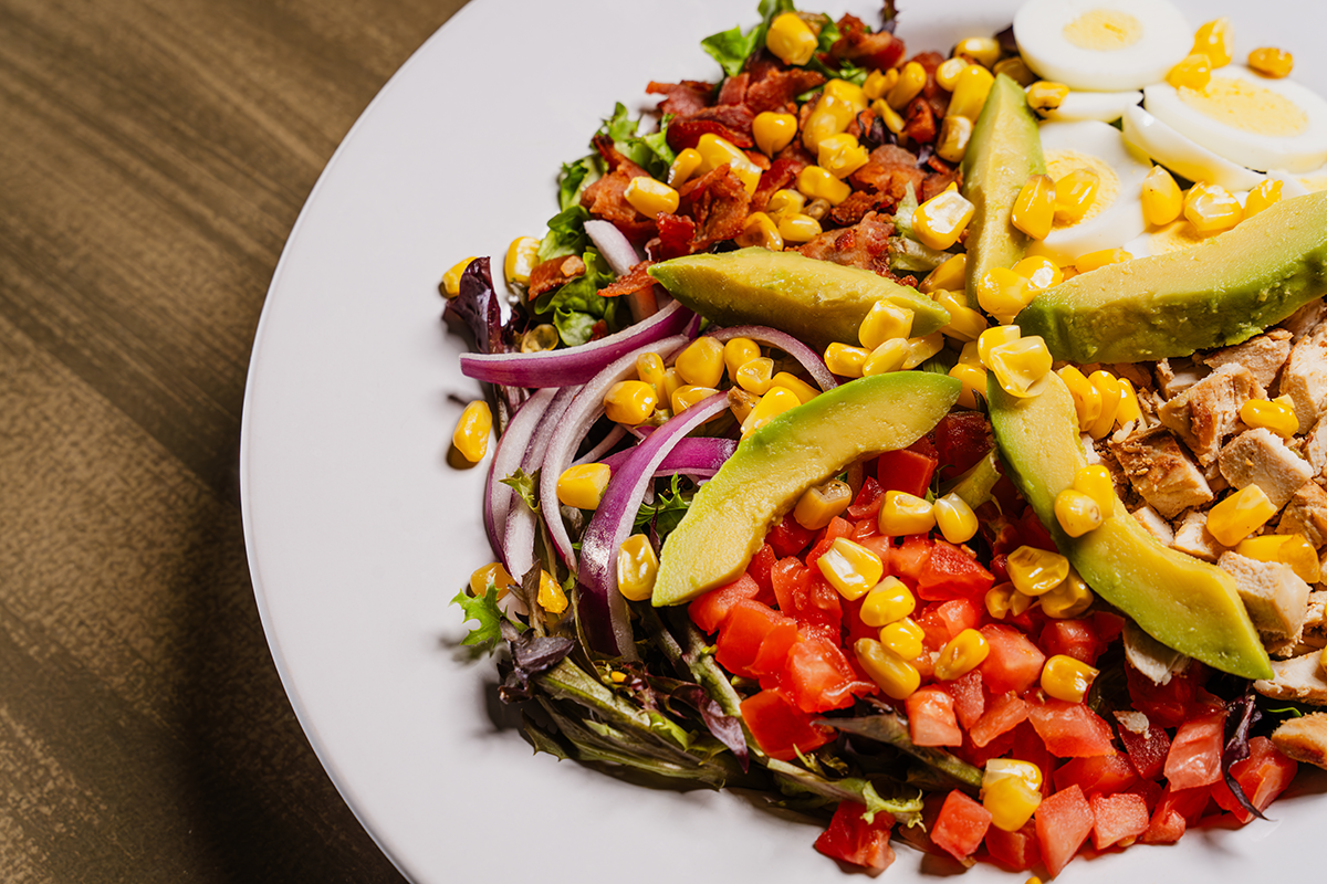 Salad on white plate with avocado, eggs, corn, tomatoes, onions, bacon, and chicken.