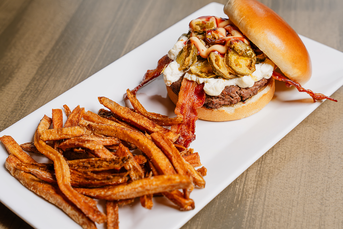 Burger with bacon, fried pickles, and sweet potato fries on a white plate.